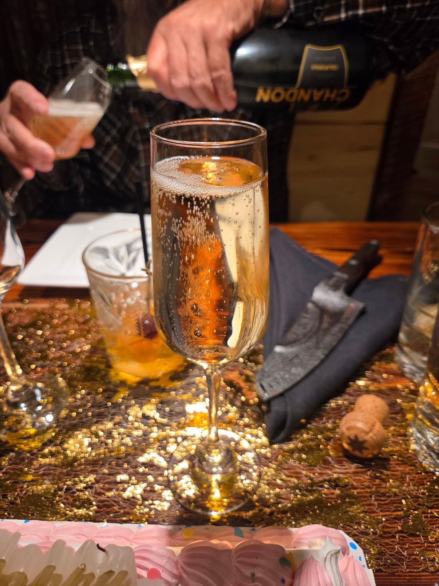 Champagne being poured into a flute beside another glass on a glittery restaurant table