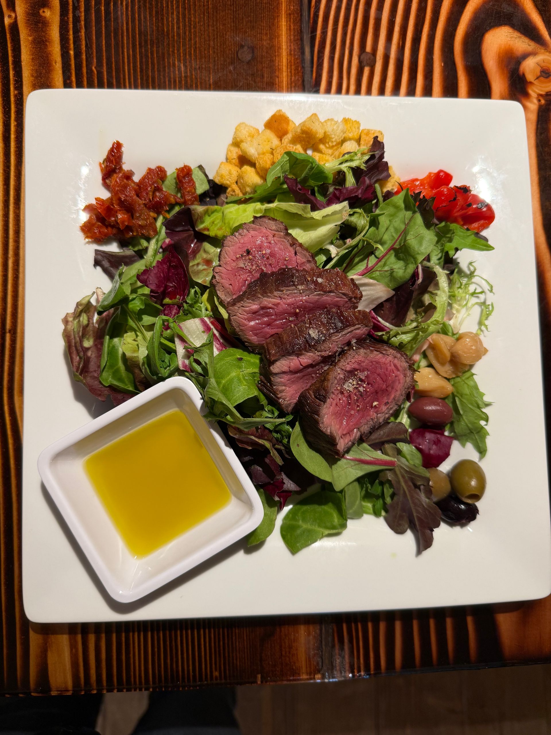 Steak salad with greens, olives, cherry tomatoes, and olive oil dressing on a square white plate