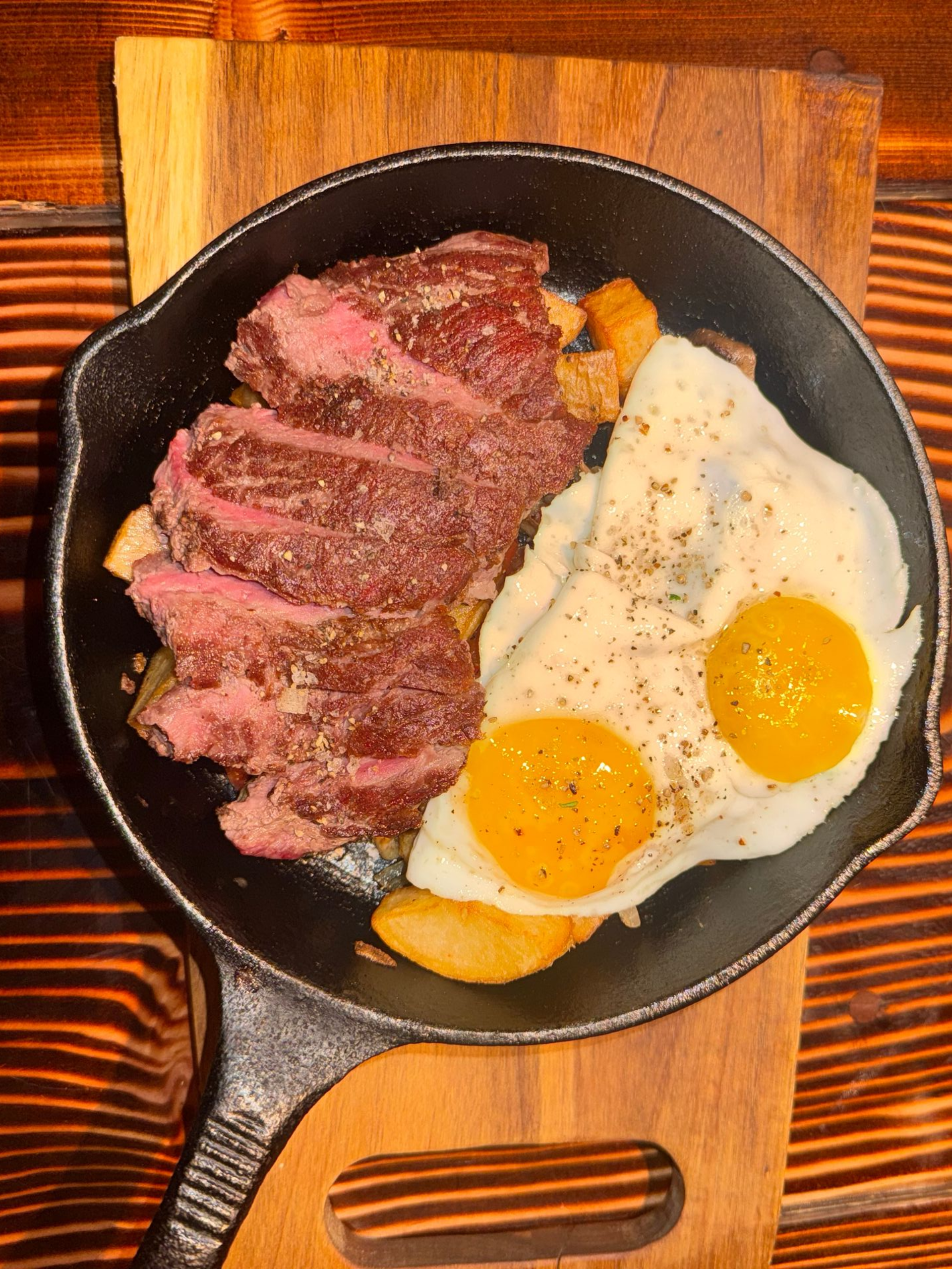 Steak with two sunny-side-up eggs in a black skillet on a wooden board