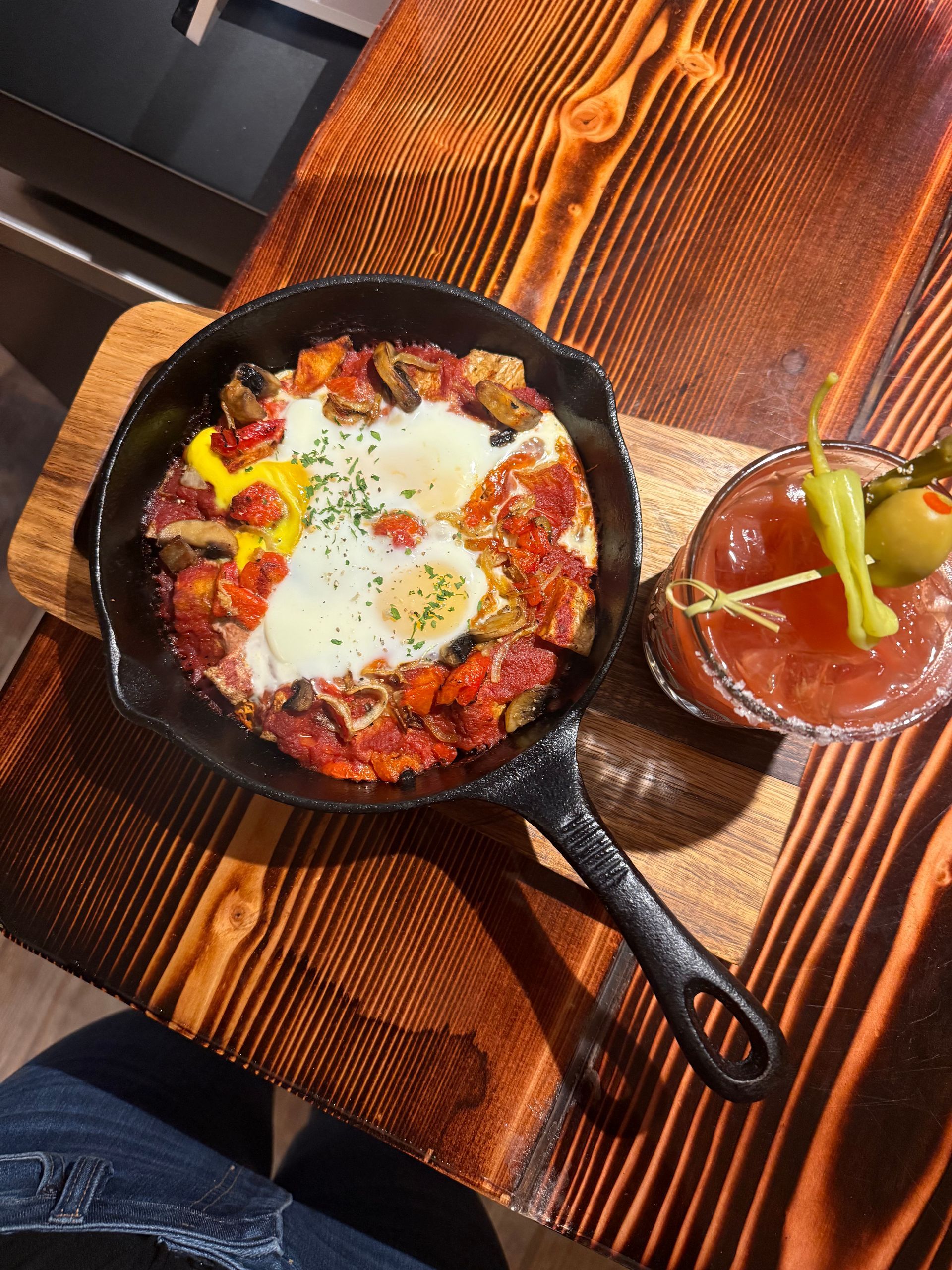 Skillet eggs with vegetables on a wooden table, beside a small bowl of red sauce and pickles.
