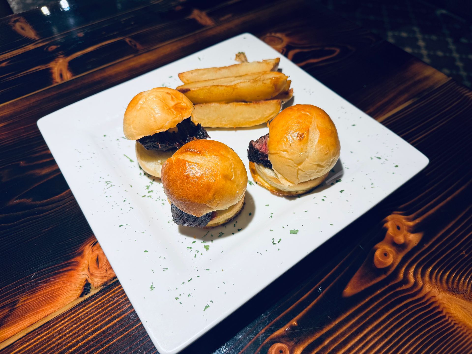 Burger sliders and fries on a white plate atop a dark wooden table