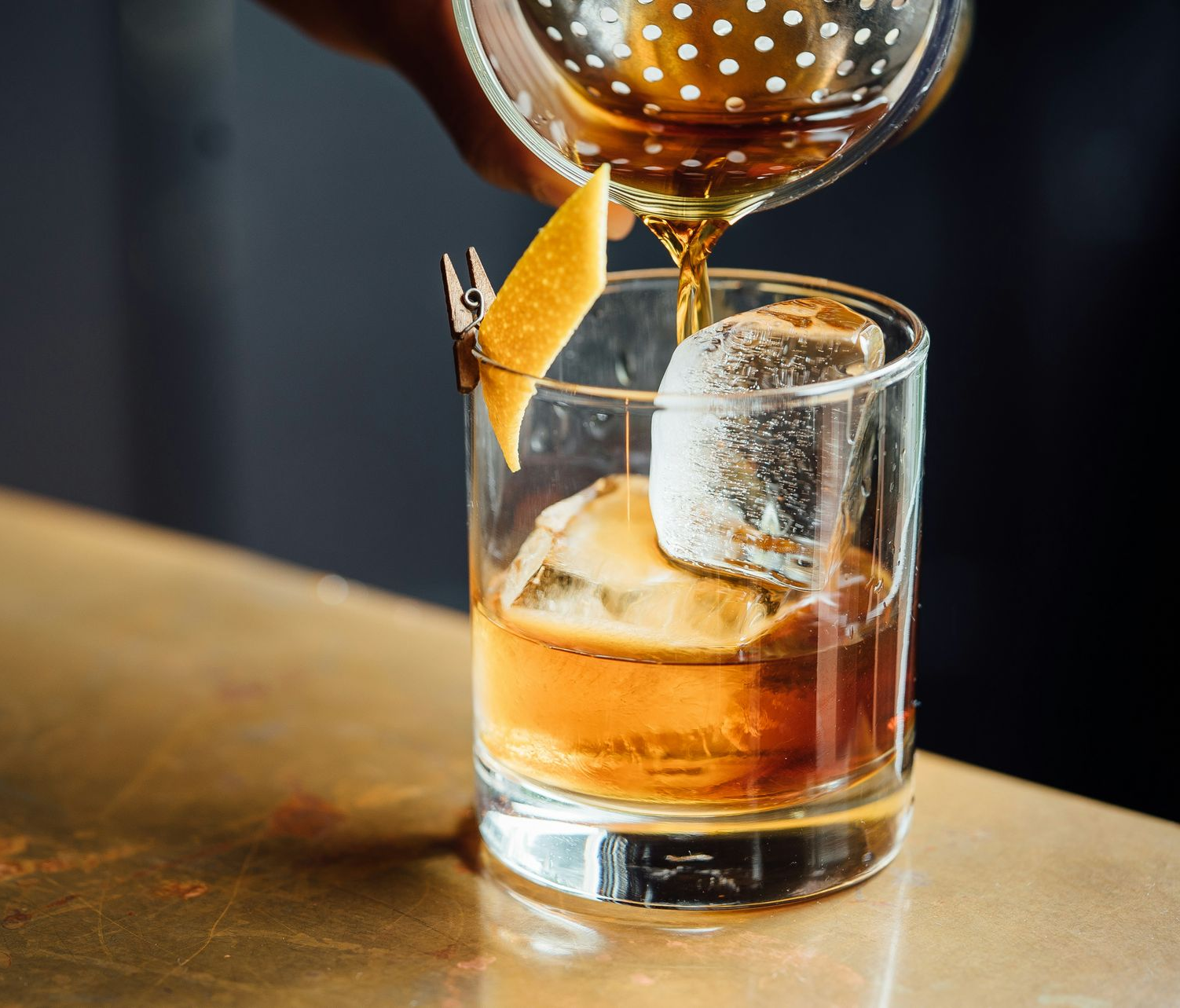 Cocktail being poured into a glass with ice on a wooden bar