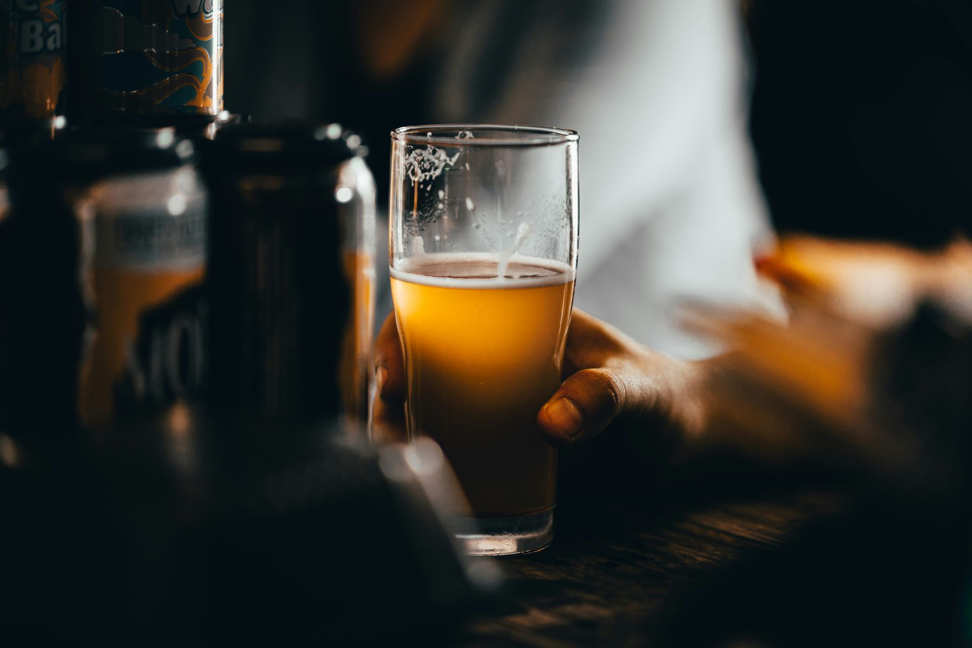 Hand holding a glass of amber beer in a dim bar, with blurred bottles in the background