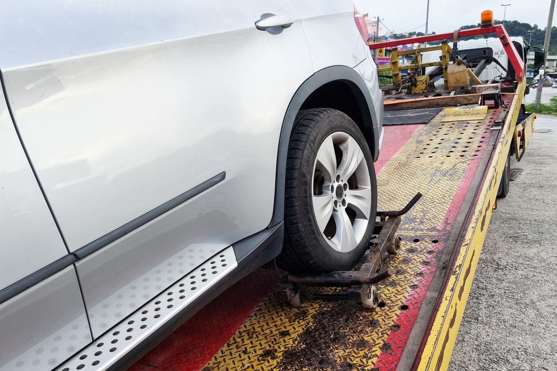 A silver vehicle sits on a tow truck bed, with its rear tire resting on a metal dolly to facilitate transport.