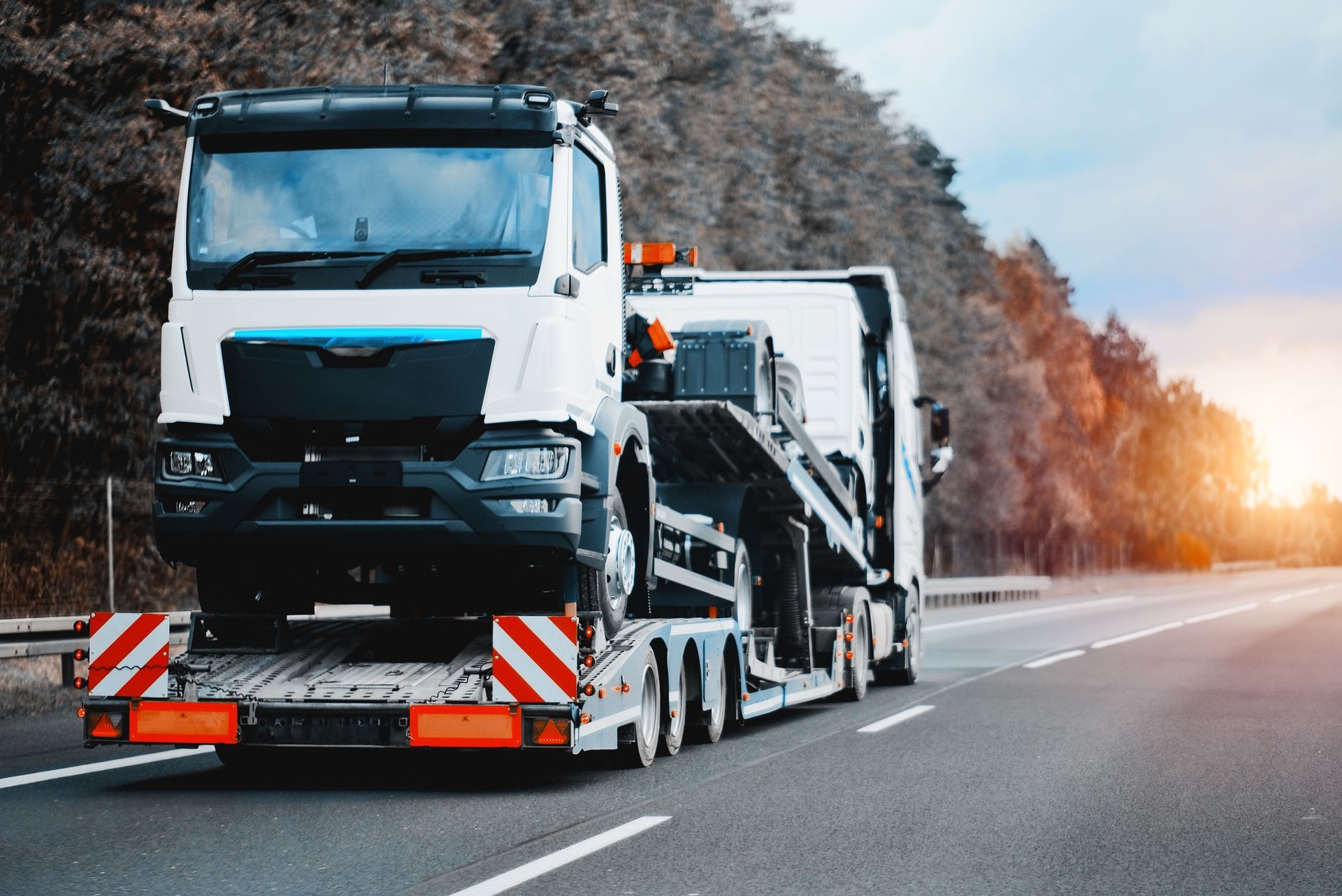 A white semi-truck carrying a smaller cab on its flatbed trailer driving along a highway during sunset.