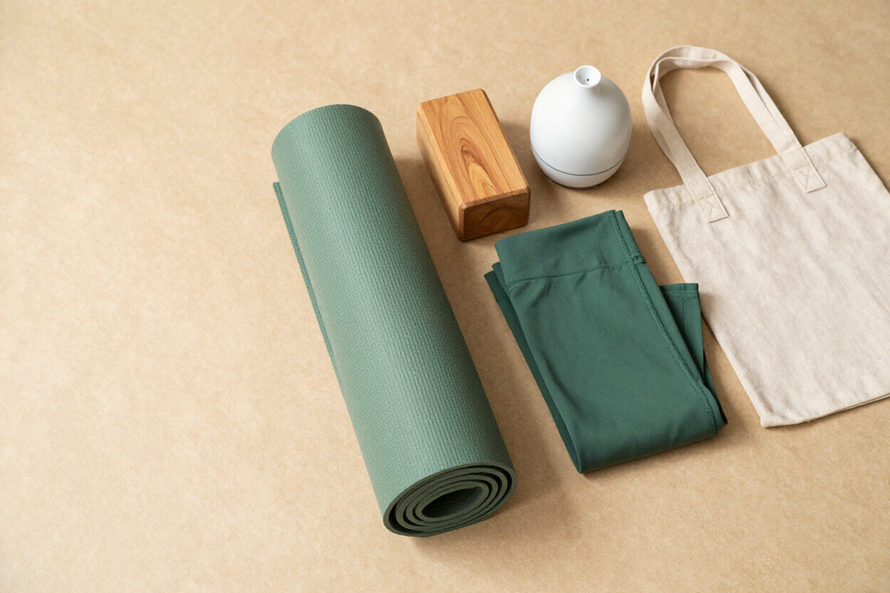 Green yoga mat, folded clothes, wooden block, white candle, and tote bag on a beige background