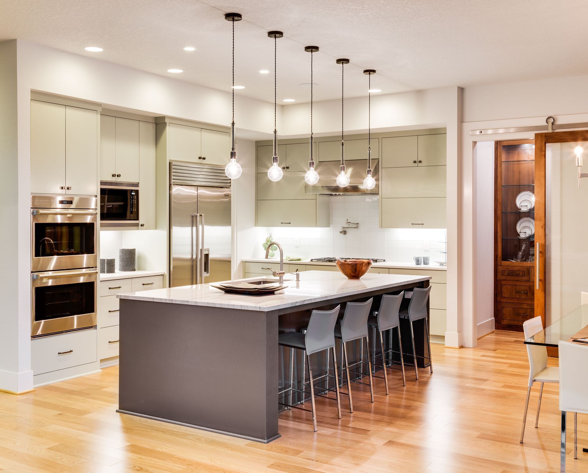 A white kitchen island with bar stools, marble countertops, and a hanging pot rack, set in a room with wood floors.