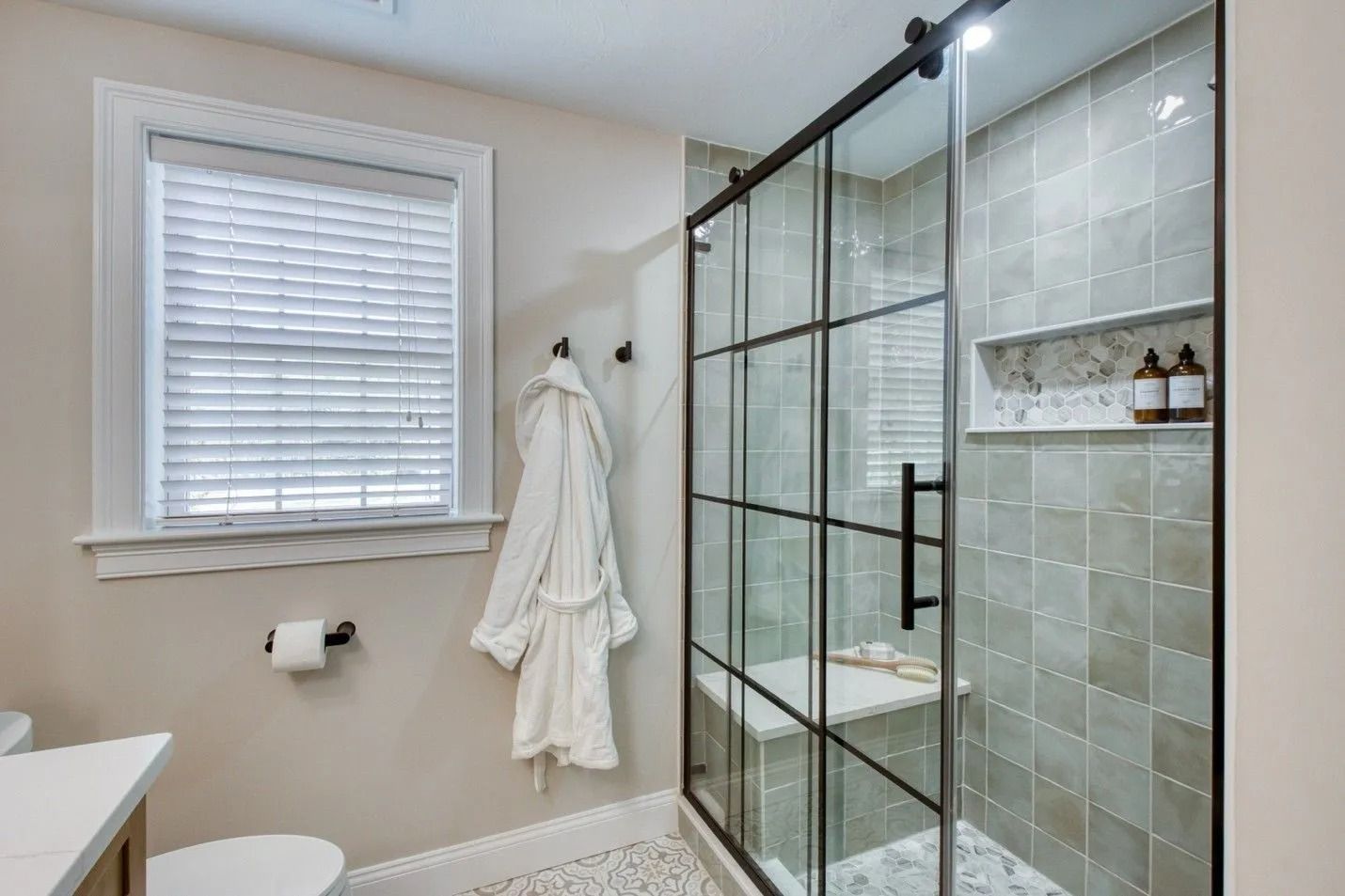 Bright bathroom with glass shower, tiled walls, window blinds, and white towels hanging on hooks