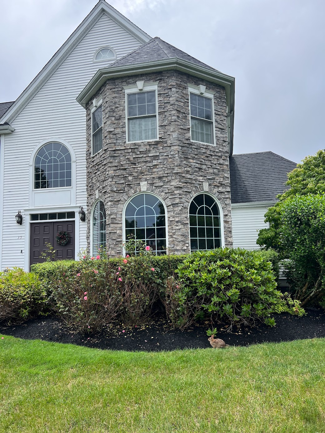 Stone and white suburban house with arched windows, trimmed hedges, and a green lawn under cloudy sky