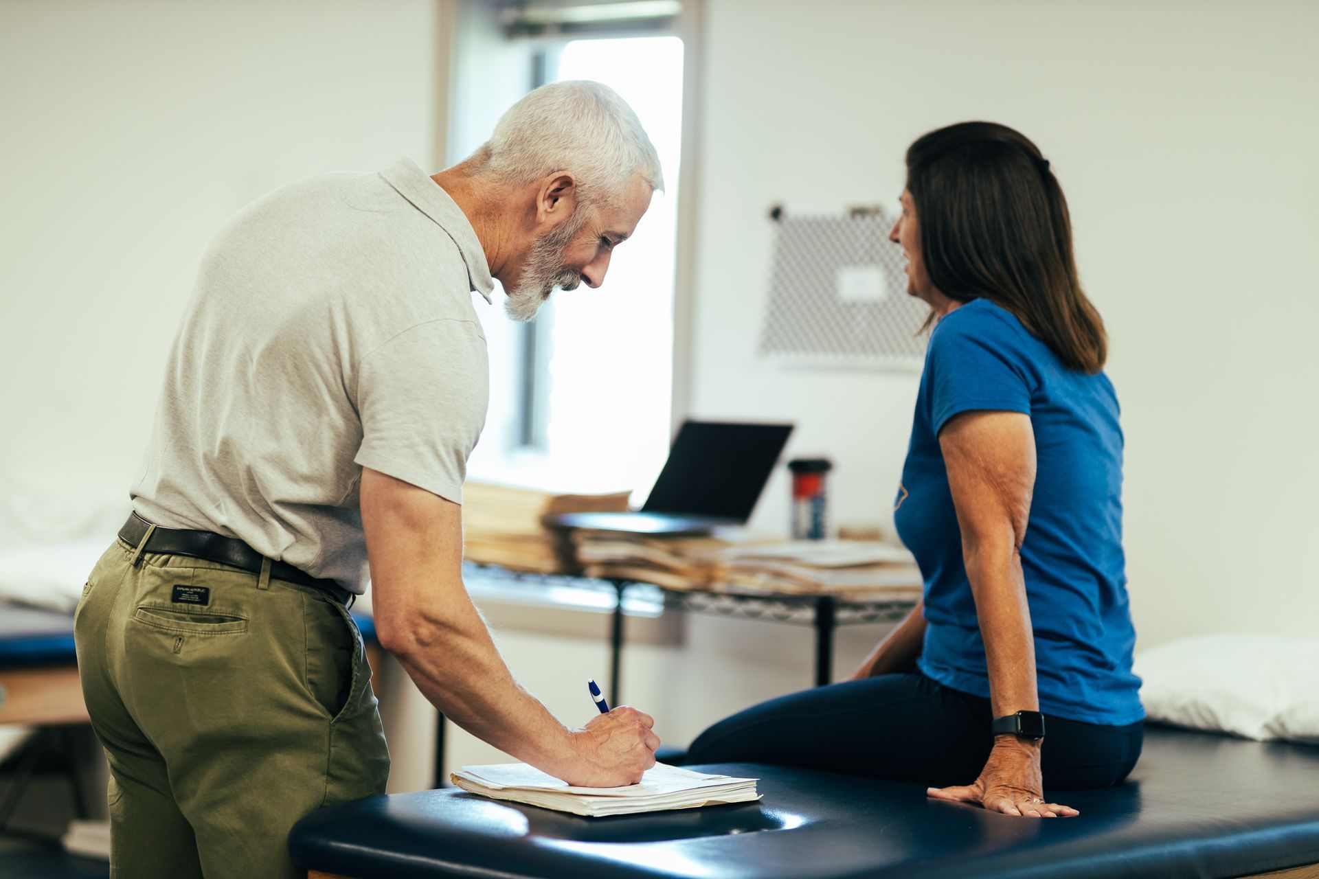 A man in khaki pants writes on a clipboard next to a woman in blue, sitting on a medical table.