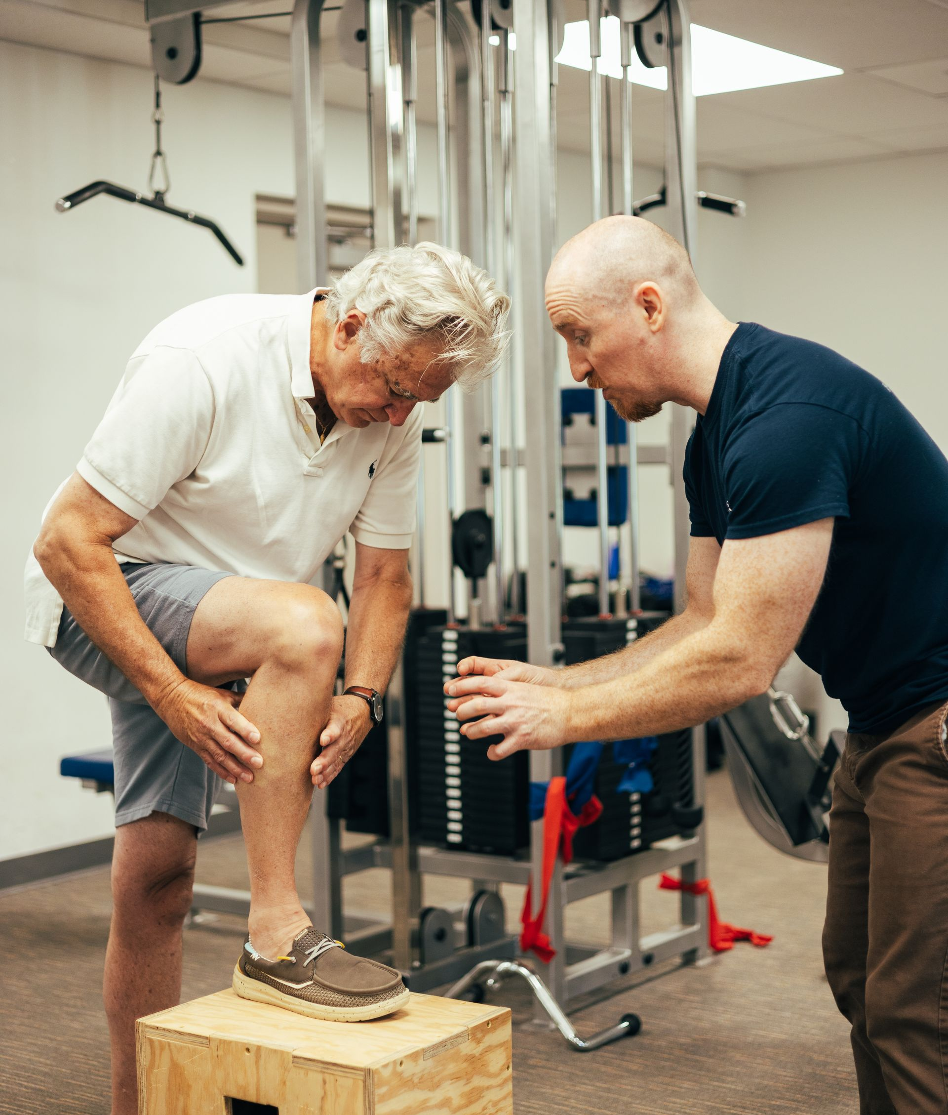 Man in shorts pointing at his leg while being assisted by a professional in a gym.