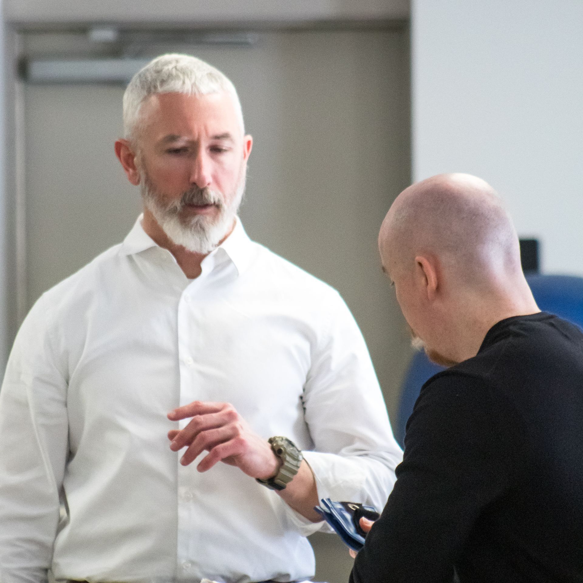 Man in white shirt gestures, talking to another man with bald head holding a device.