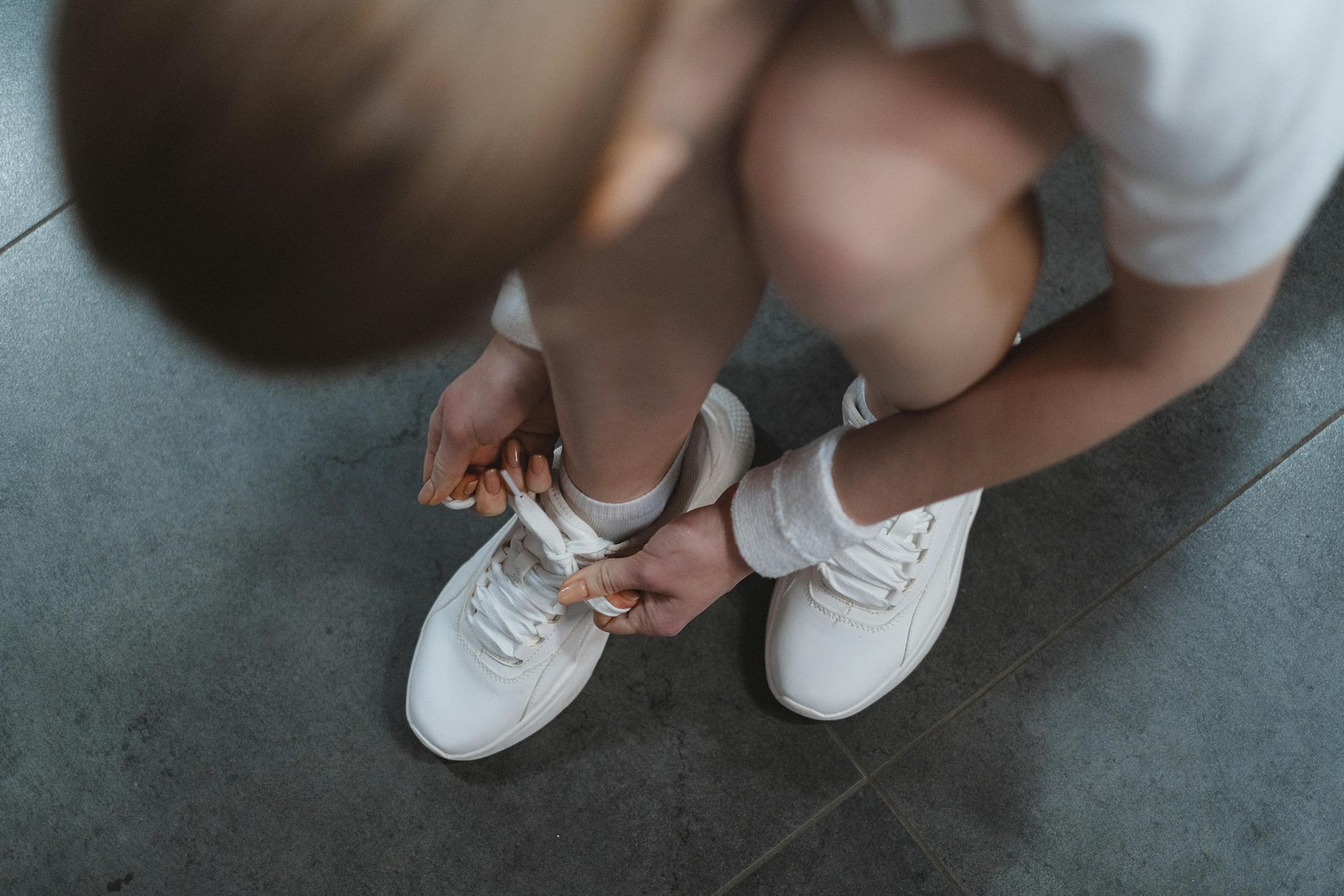 Person tying white shoelaces, wearing white sneakers and wristband, on a grey tiled floor.