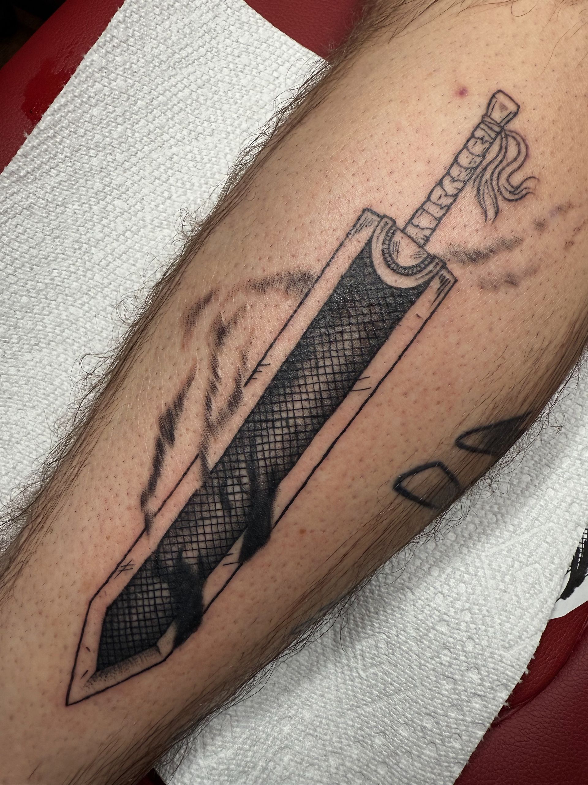 Black and gray tattoo of a pencil-like object on a forearm, shown on a white towel with a red background.