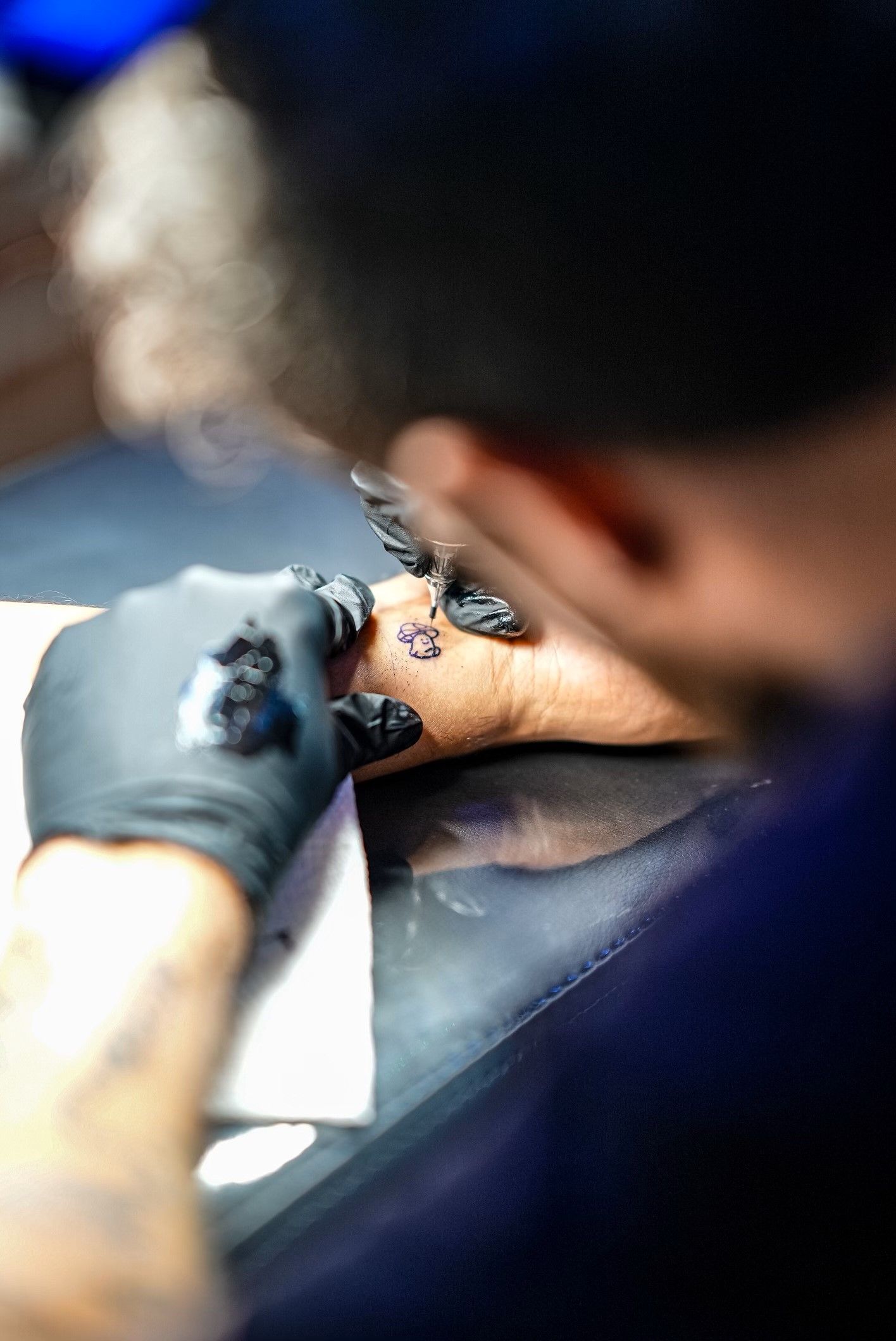 Gloved hand tattooing ink onto a person’s wrist on a tattoo chair