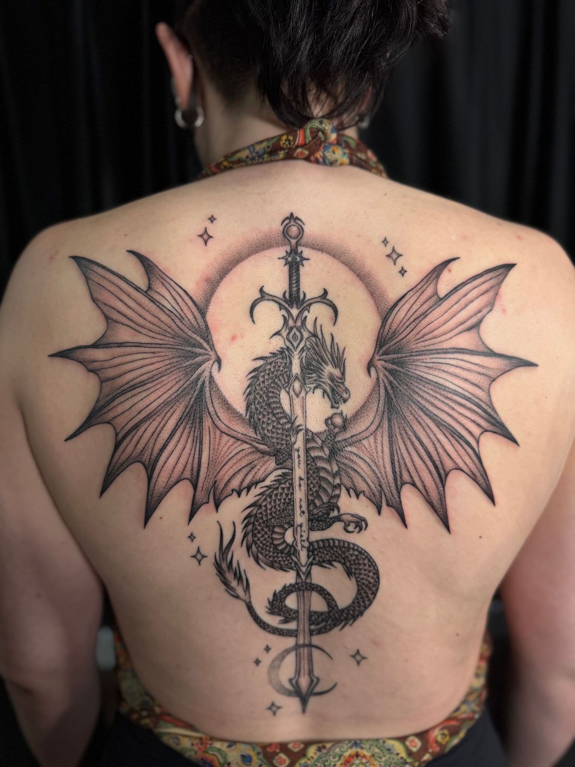 Back tattoo of a winged serpent-like sword design in black and red ink on a person's upper back