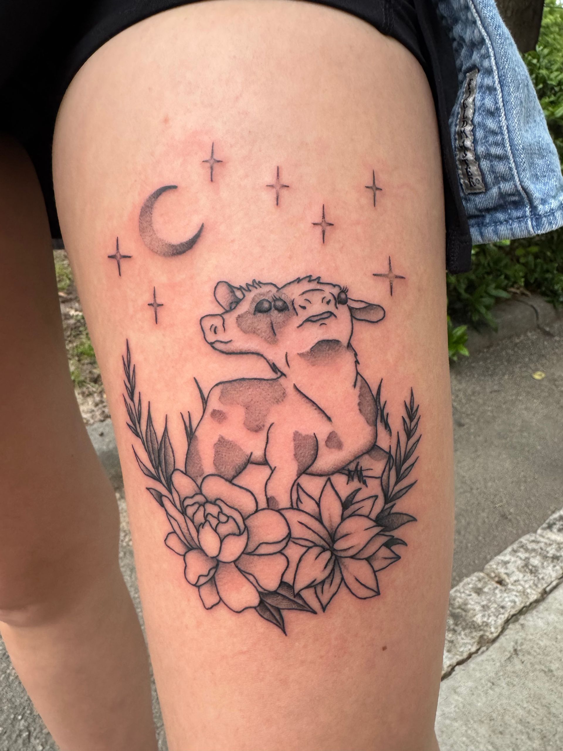 Black line-art tattoo of a cat on flowers with crescent moon and stars on a thigh