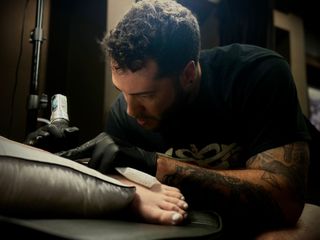 Man tattooing a client’s arm at a tattoo studio, leaning over the work with focused expression