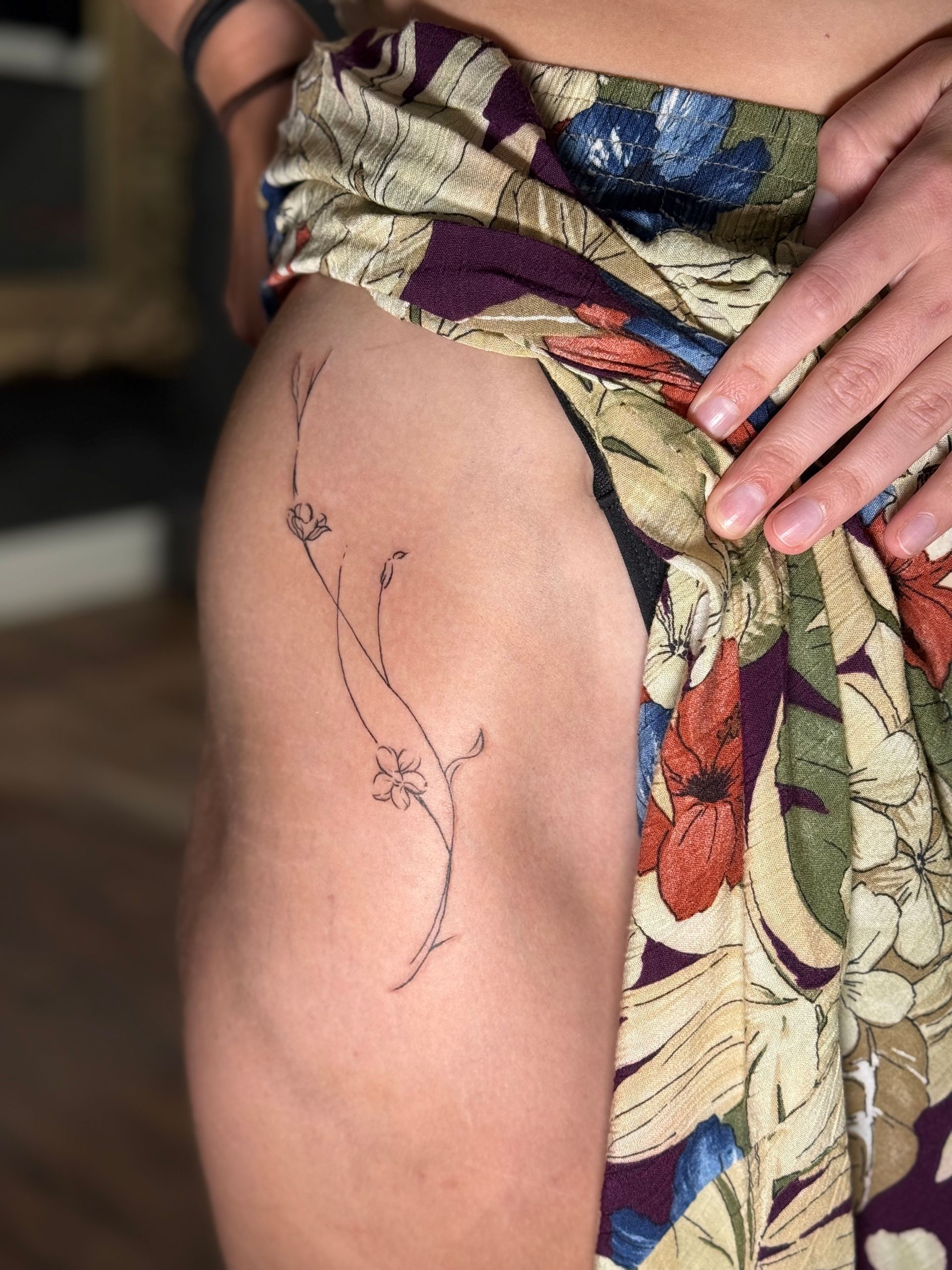 Person showing a thigh with a fine black floral line tattoo and a patterned wrap skirt