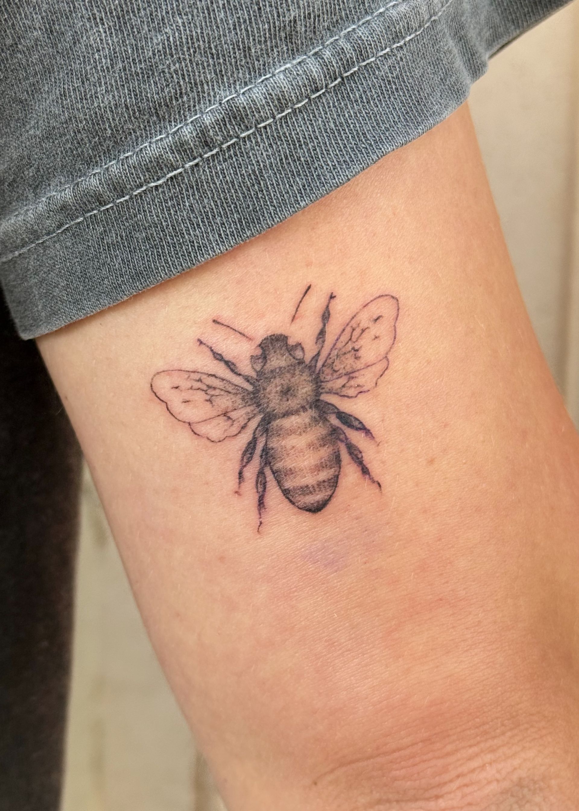 Bee tattoo on a forearm with purple-black ink, shown near the sleeve cuff.