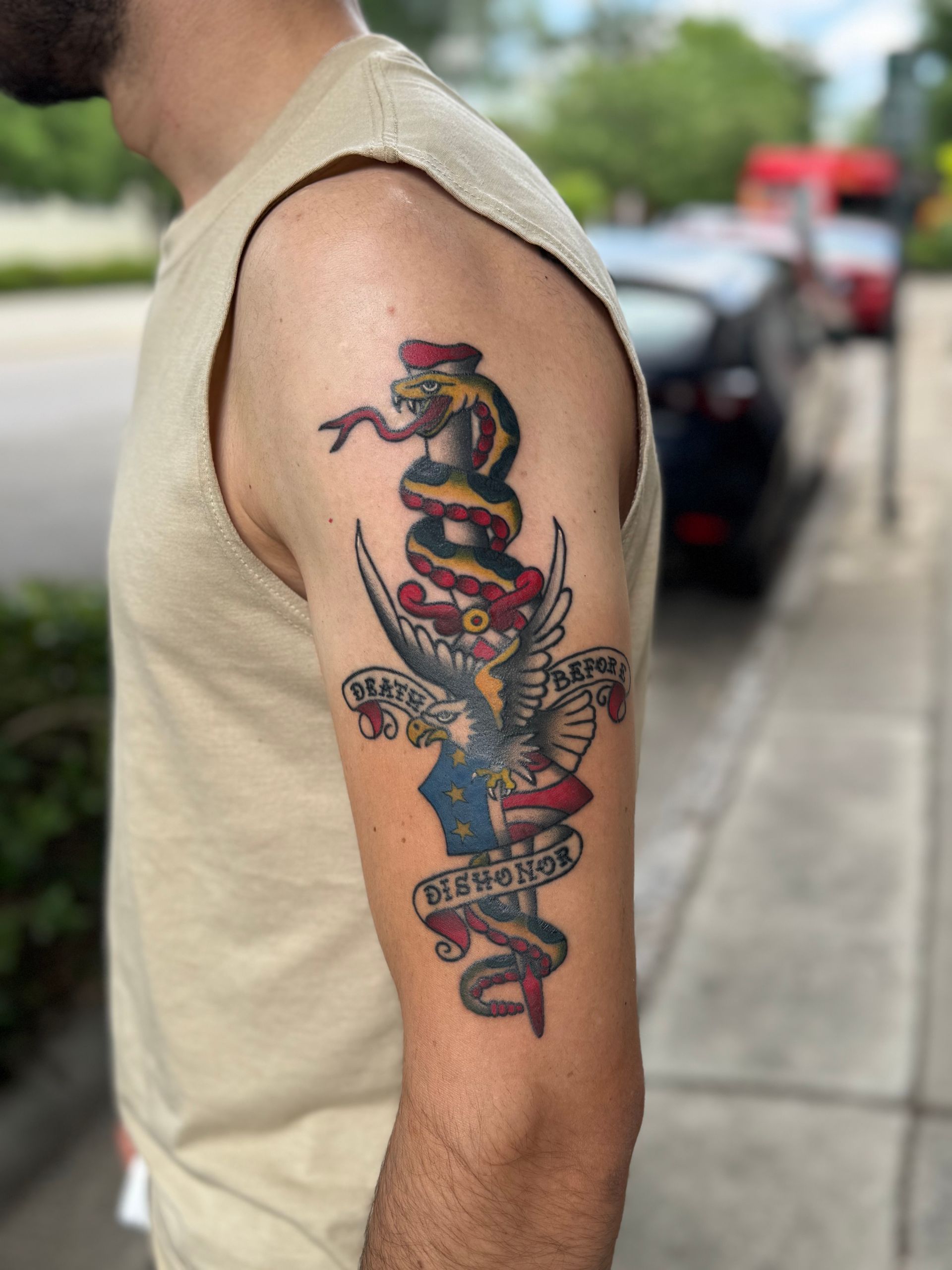 Arm tattoo of a colorful snake entwined with a dagger on a person’s upper arm outdoors