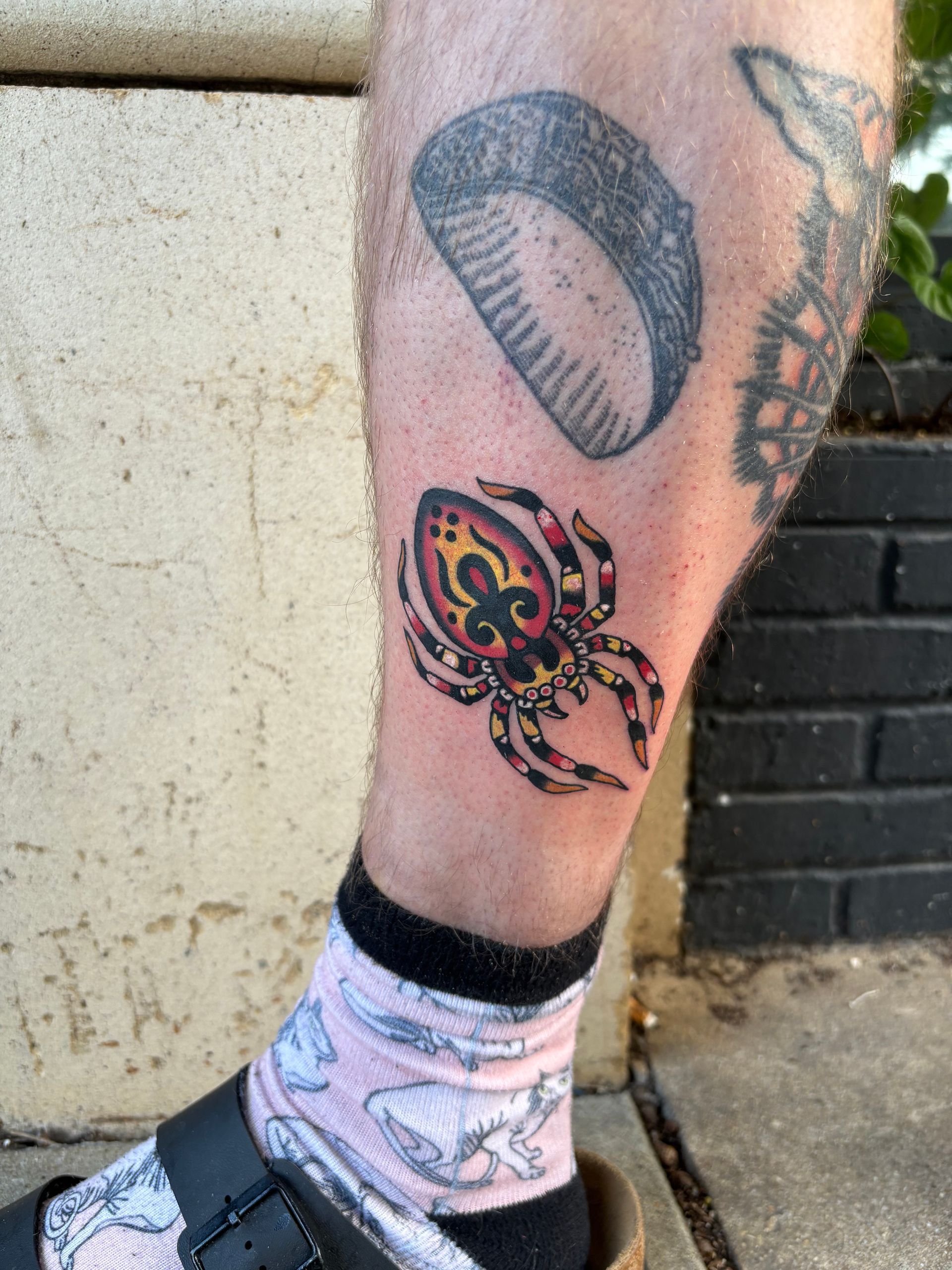 Tattooed lower leg with a colorful spider tattoo and black shoes on a concrete path