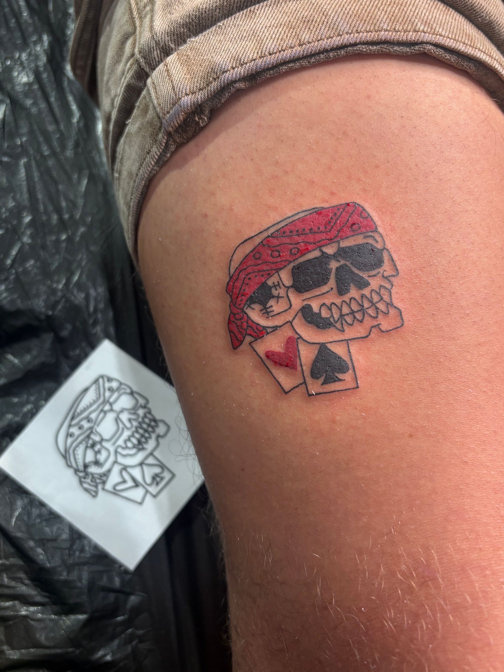 Small skull tattoo with red hat on upper arm, beside a black-and-white tattoo stencil.