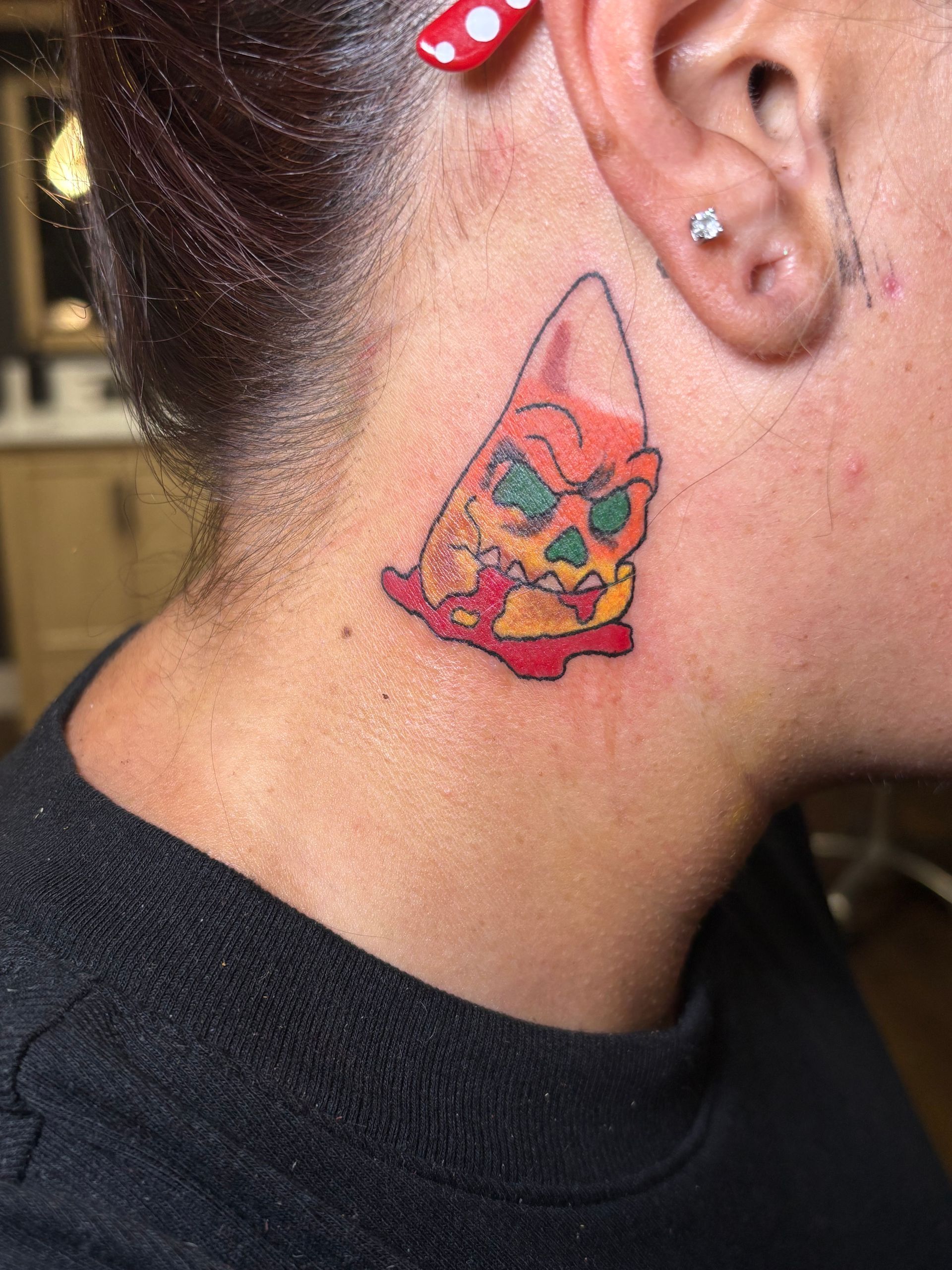 Small colorful tattoo of a cartoon cat on the side of a neck, near an ear, with a red hair clip.