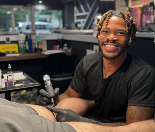 Tattoo artist smiling while tattooing a client in a shop, wearing black gloves and holding a tattoo machine