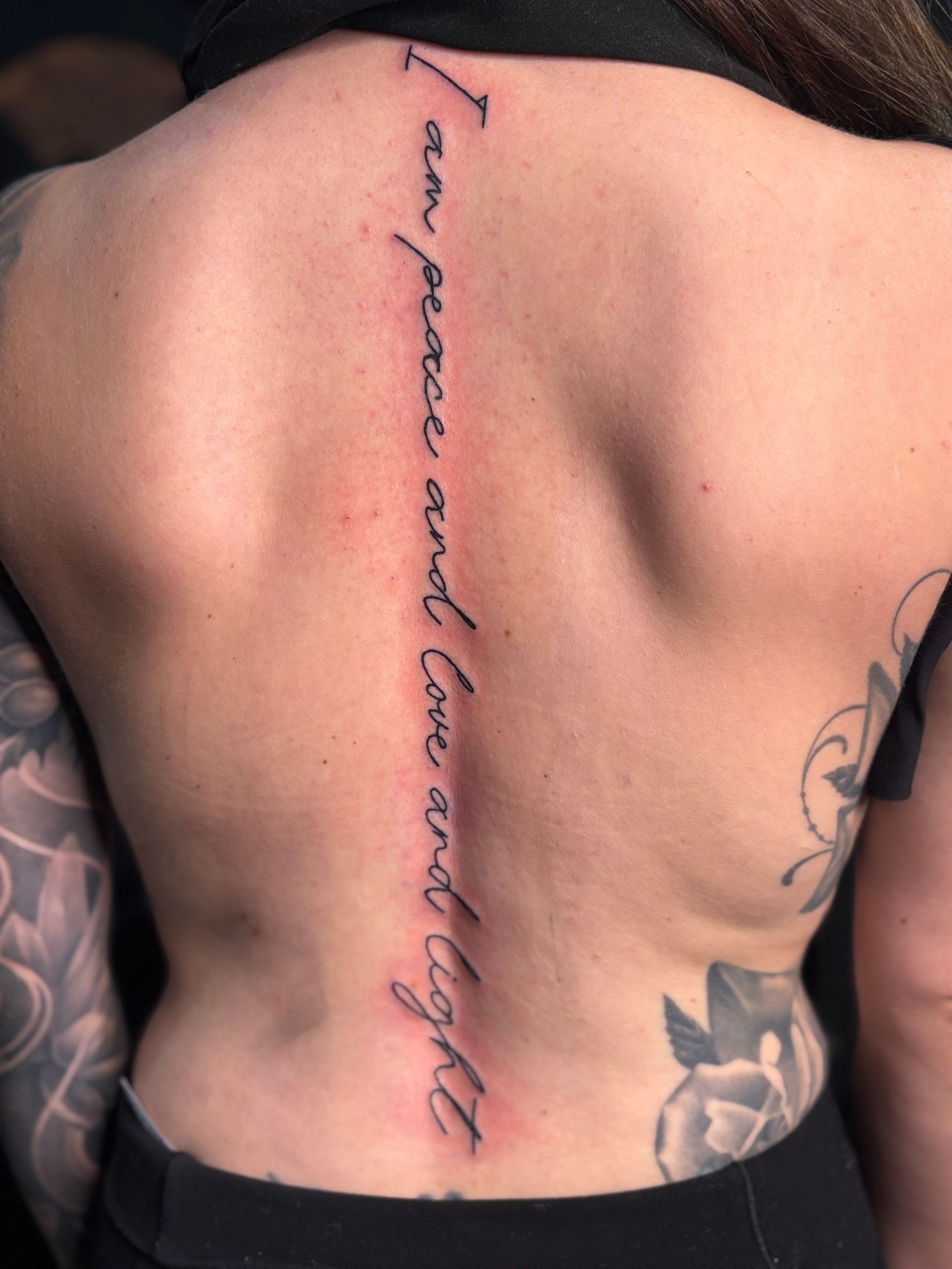 Woman’s bare back with a vertical cursive tattoo along the spine and floral tattoos on the side.