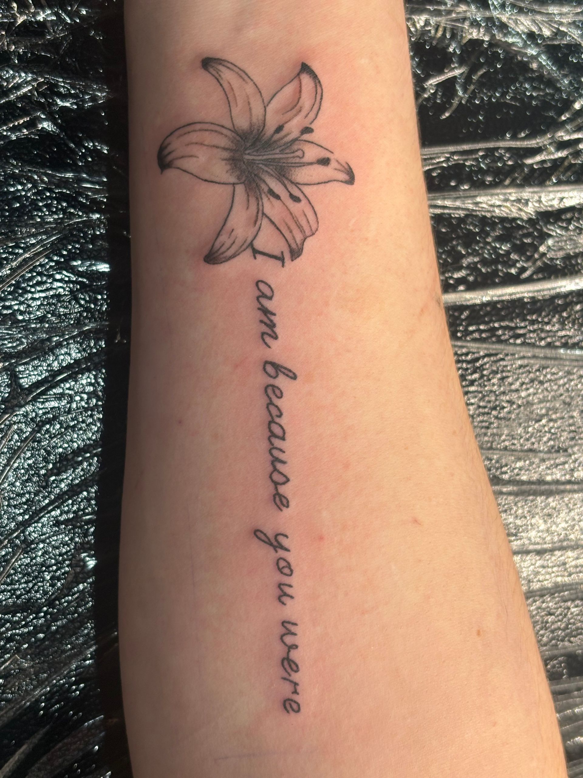 Forearm tattoo of a flower with the cursive phrase “I am because you were.”