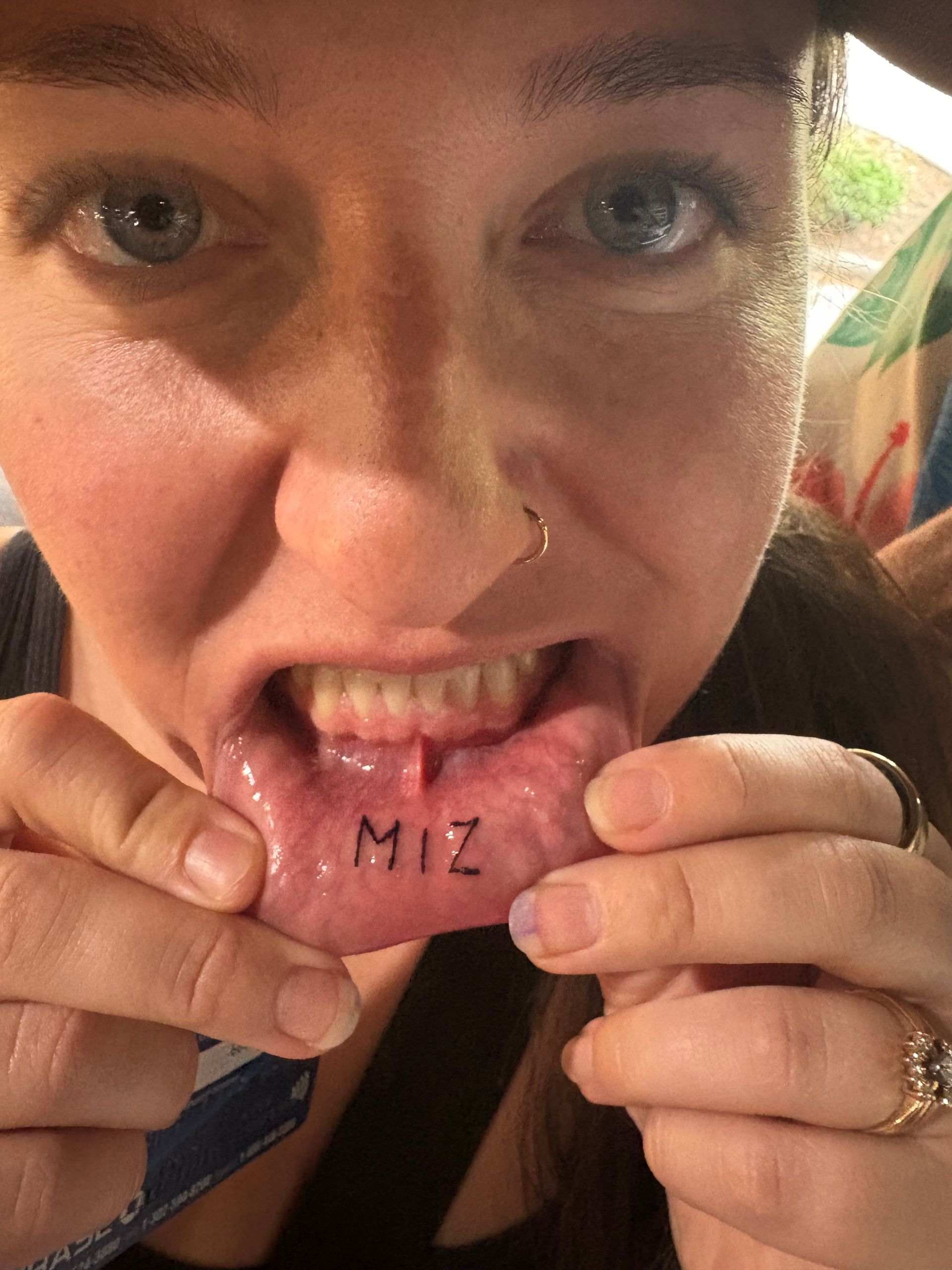 Close-up of a person biting a pink tag labeled “MIZ,” with a nose ring and rings visible.