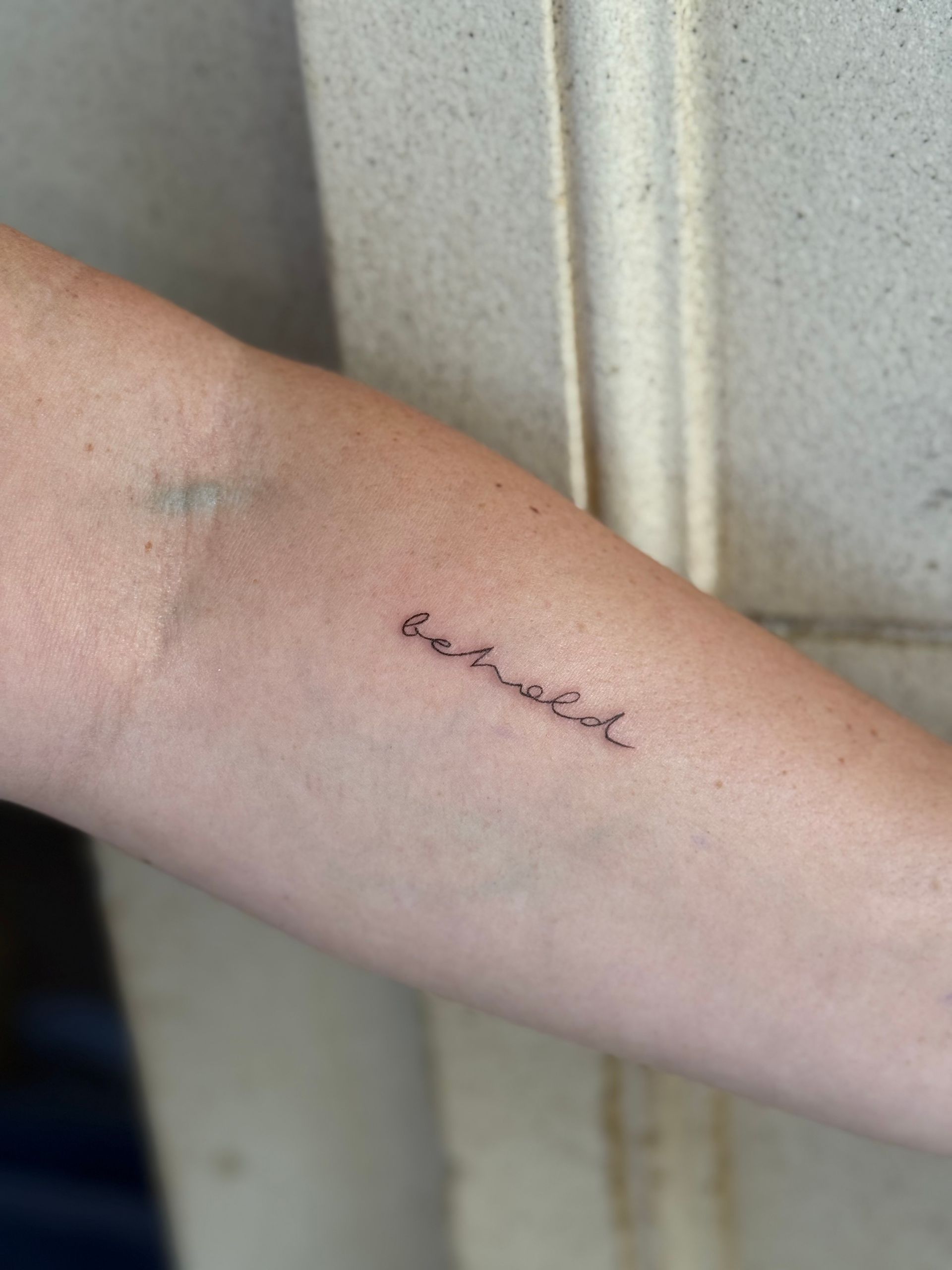 Forearm with a small cursive tattoo near the elbow, against a light-colored wall.