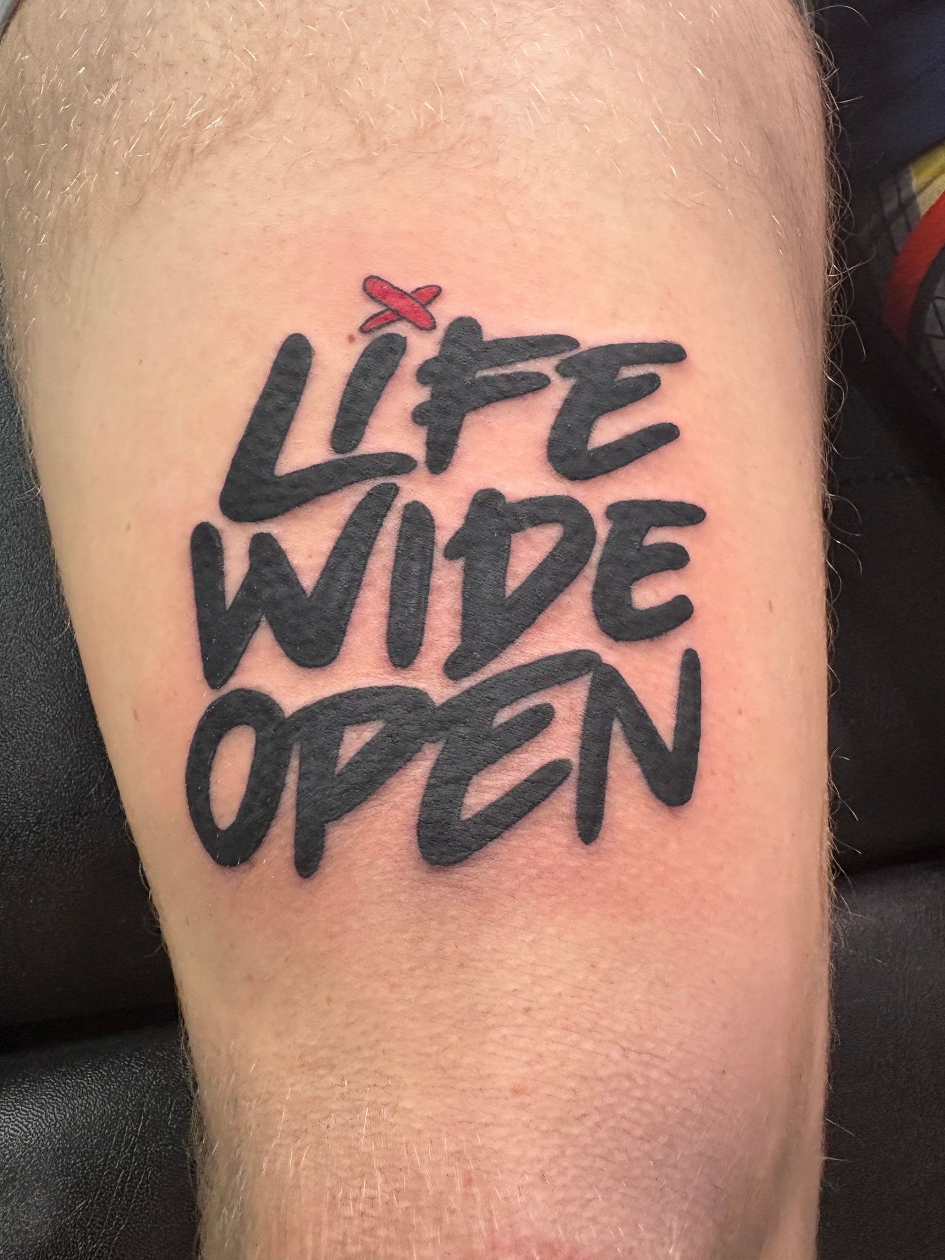 Close-up of a tattoo reading “LIFE WIDE OPEN” in black with a small red bird above it