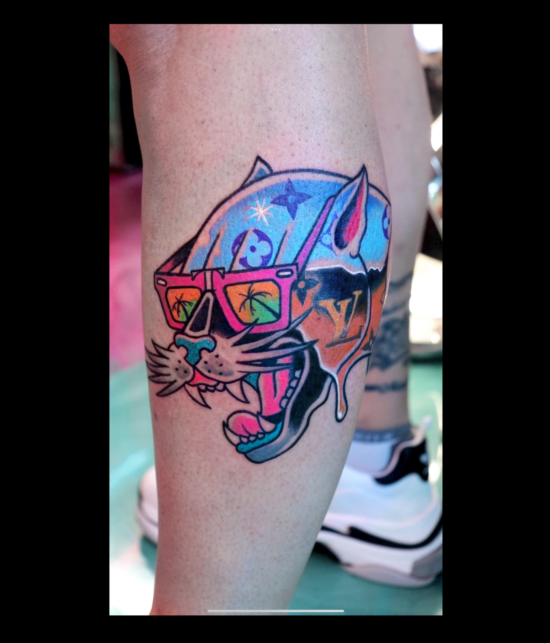 Colorful cat tattoo on a calf, wearing pink sunglasses and a blue cap, mouth open in a playful grin.
