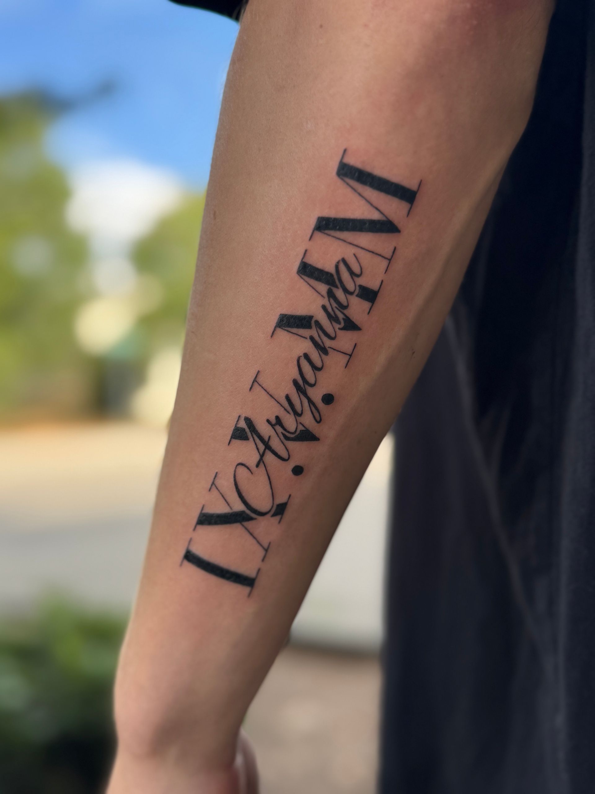Close-up of a forearm with a black vertical tattoo against a blurred outdoor background