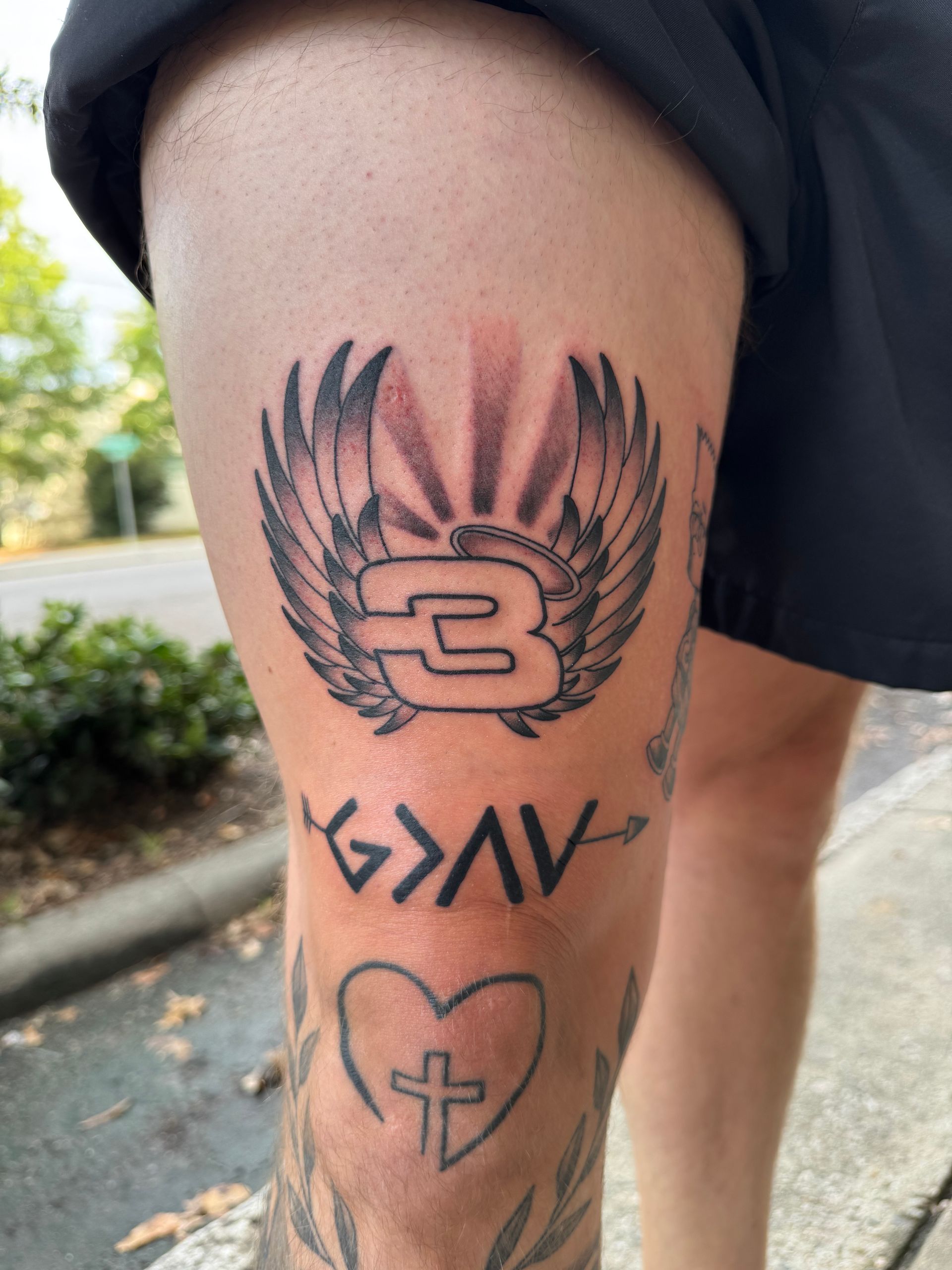 Close-up of an arm with three tattoos, including a winged letter B, symbols, and a heart, outdoors.