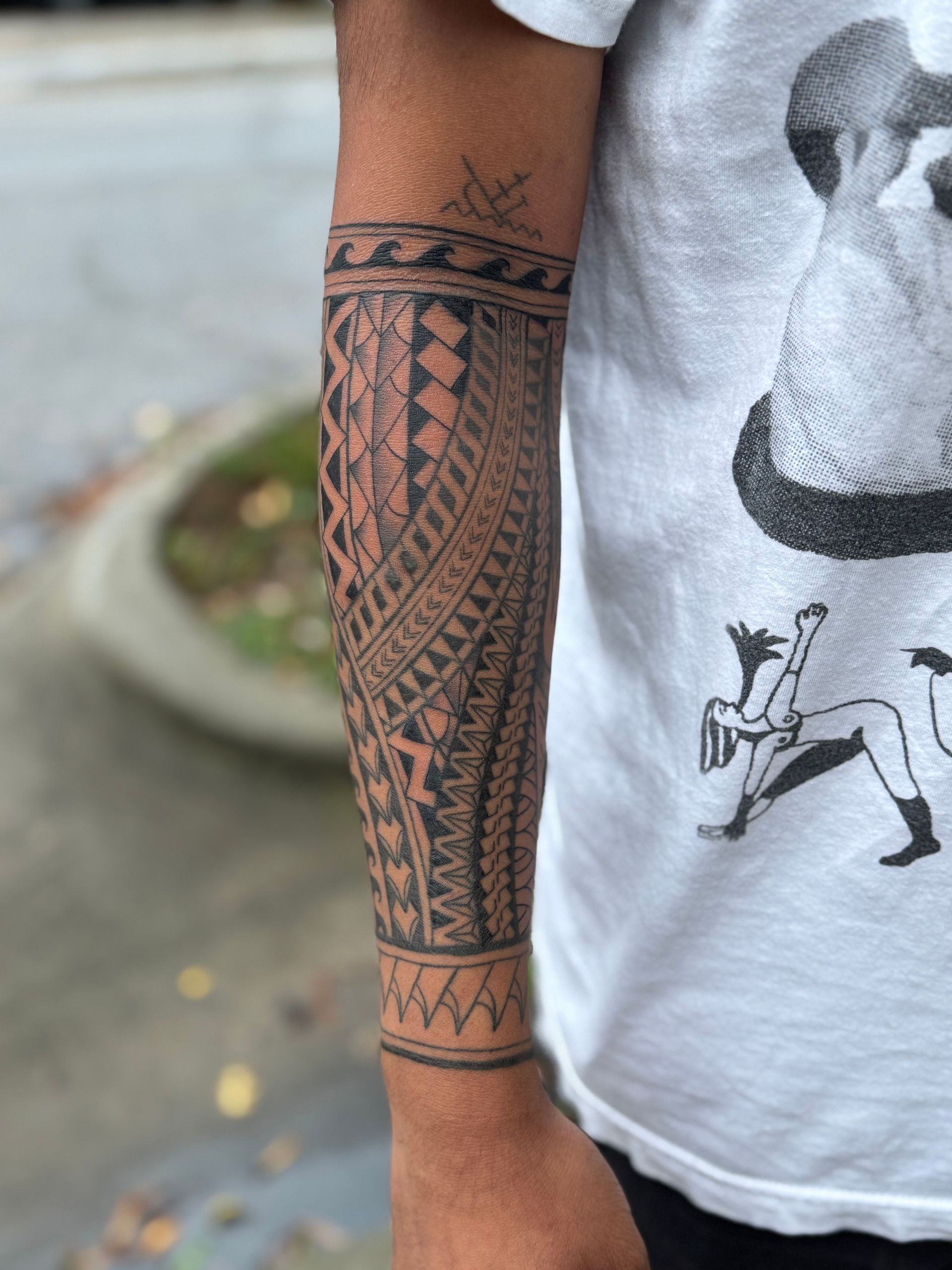 Close-up of an arm with a geometric black tattoo sleeve and bracelet pattern next to a white graphic T-shirt