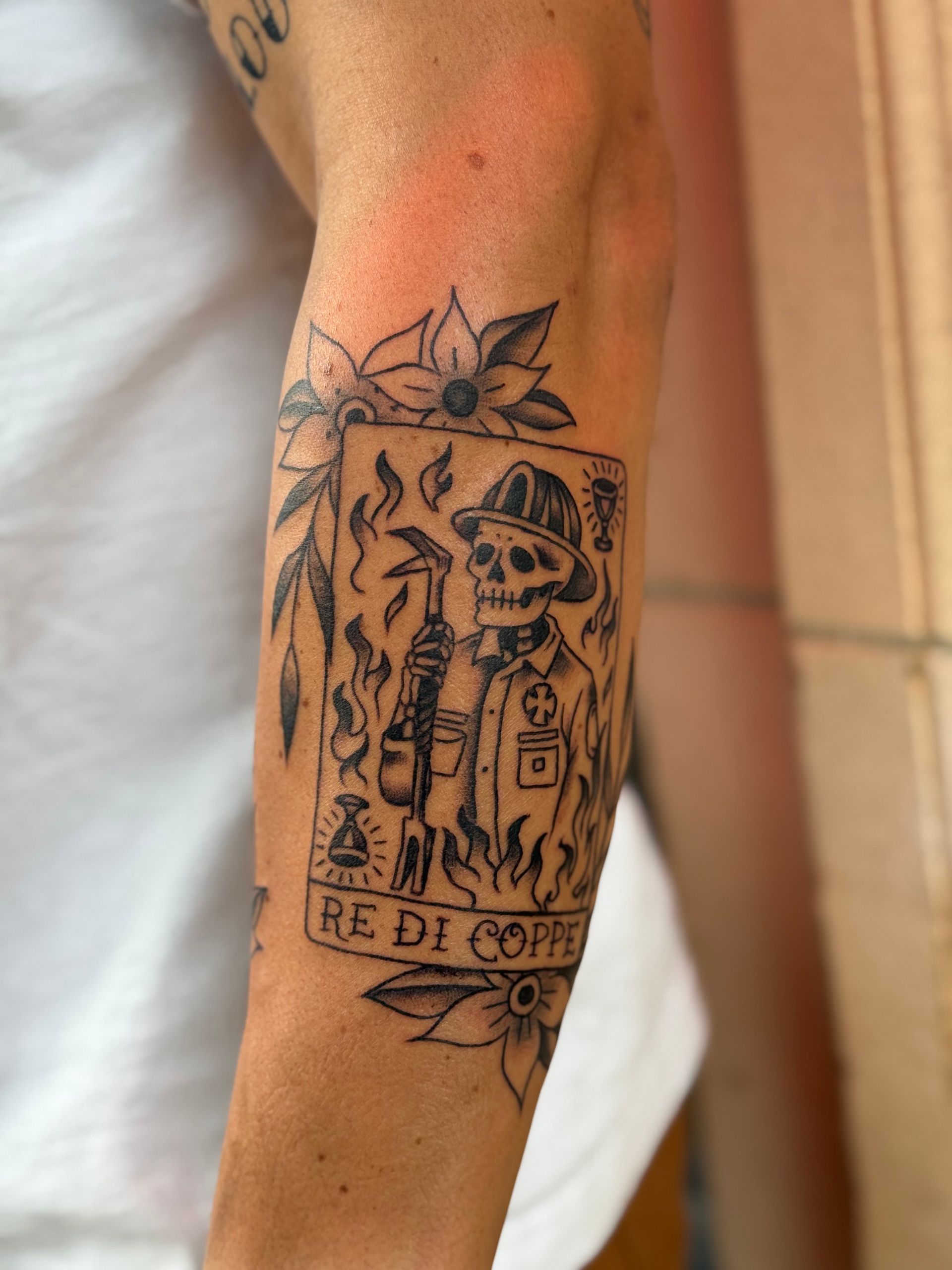 Black linework tattoo on a forearm showing a skull and skeleton figures inside a framed design.