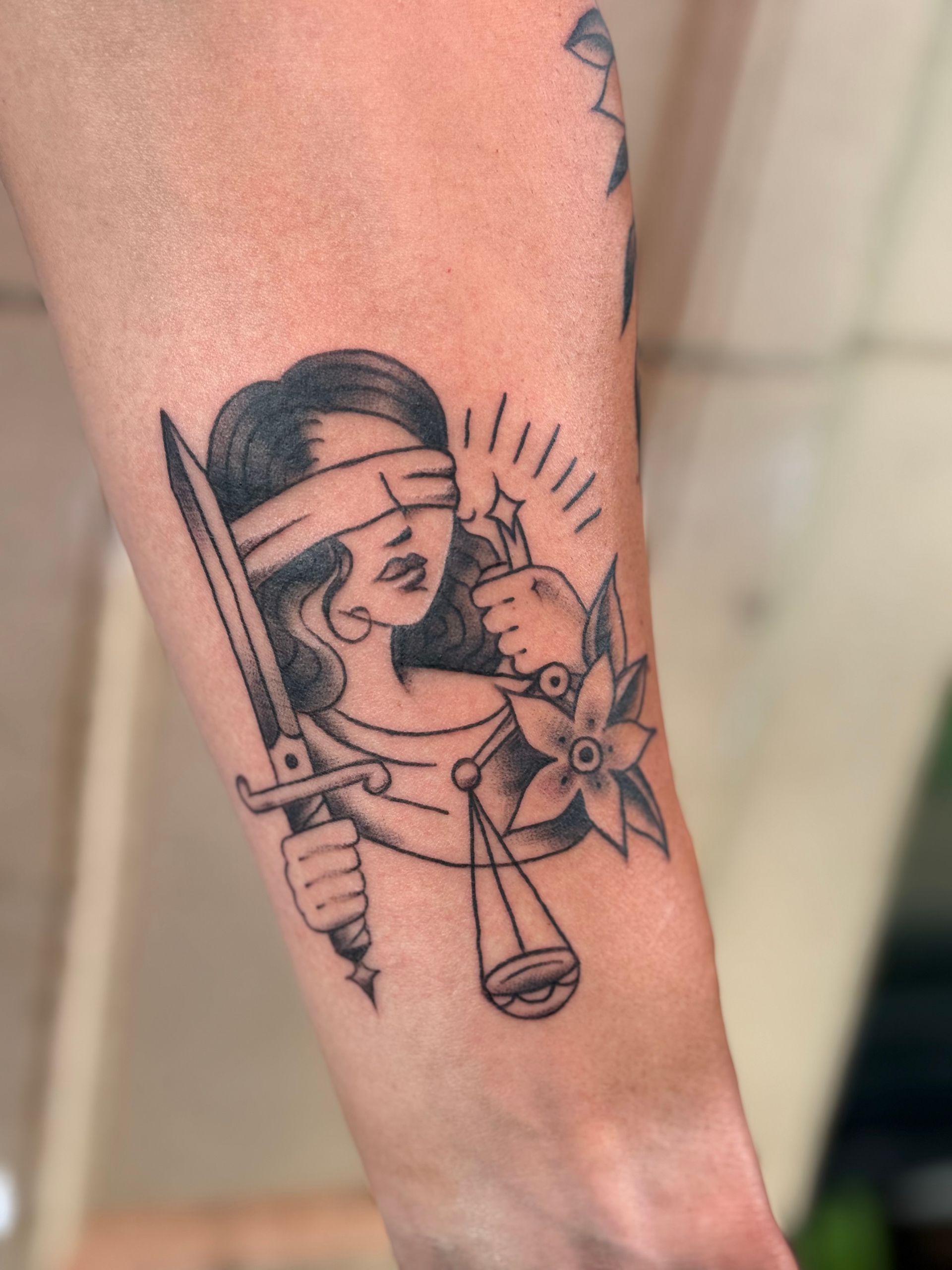 Black line-art tattoo of Lady Justice holding a sword and scales on a forearm.