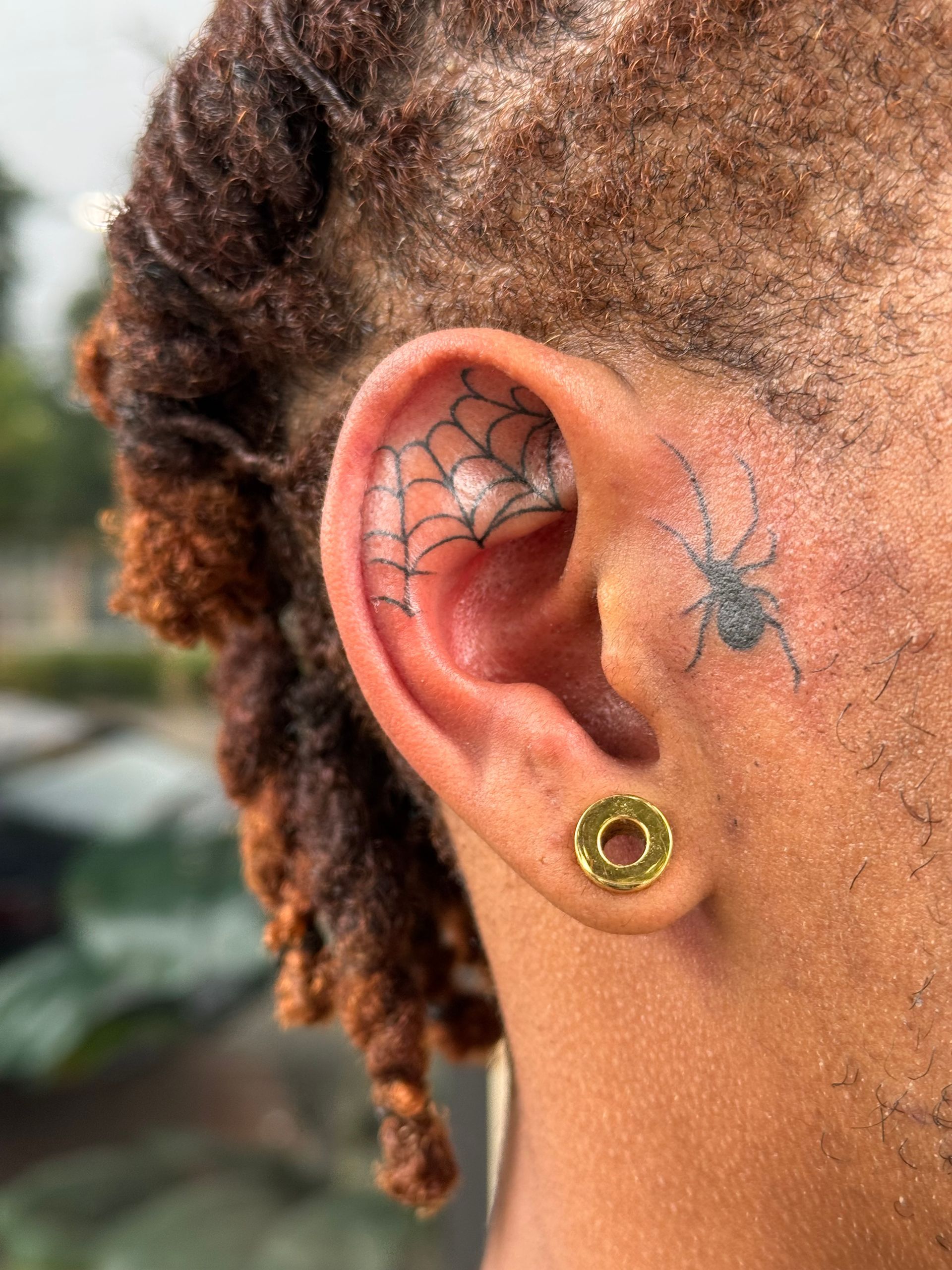 Close-up of a person’s ear with spiderweb and spider tattoos and a gold ear gauge