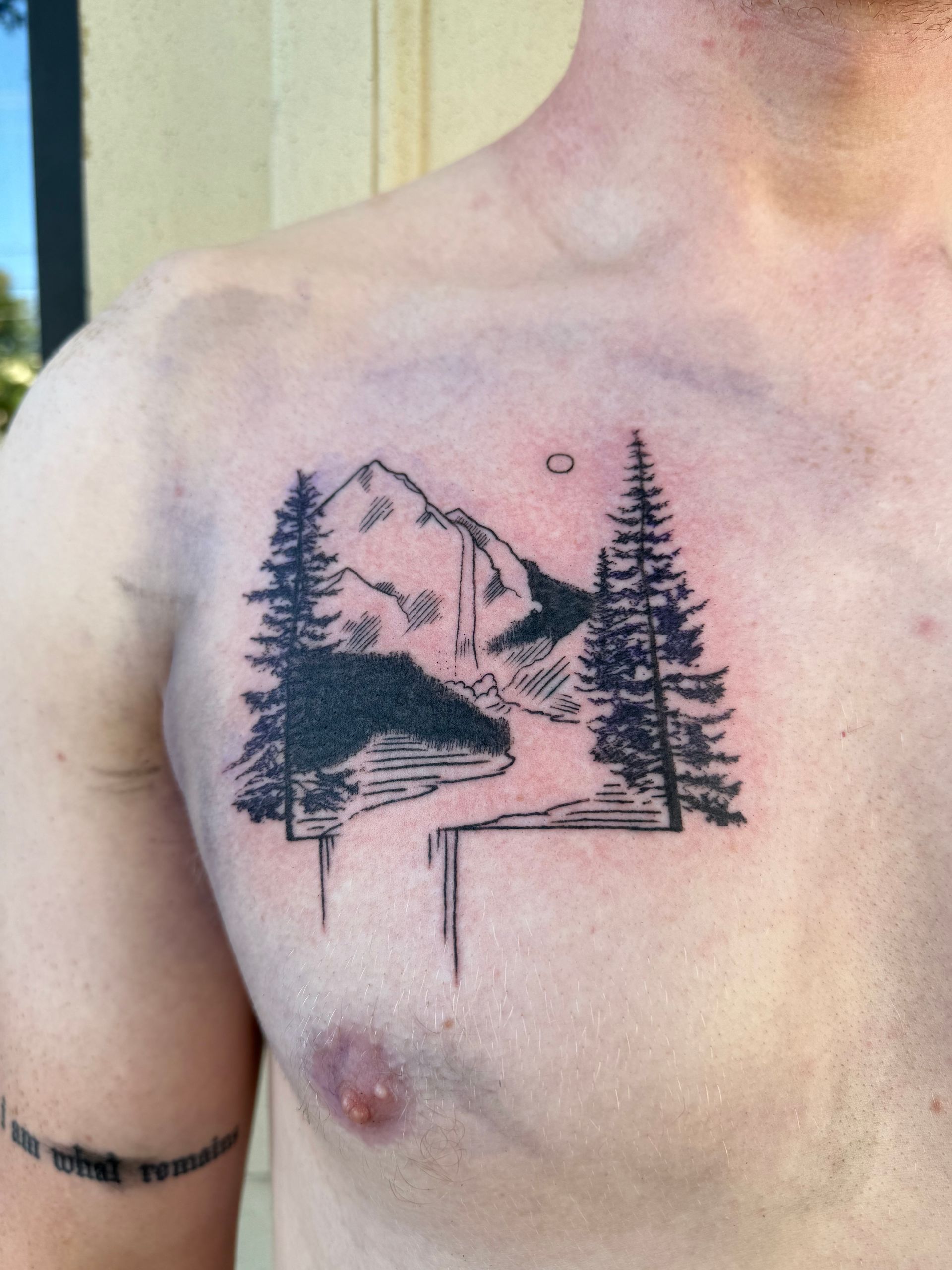 Chest tattoo of a mountain, pine trees, and a river in black ink