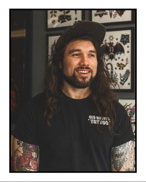 Smiling man with long hair and tattoos, wearing a black cap and T-shirt, standing indoors