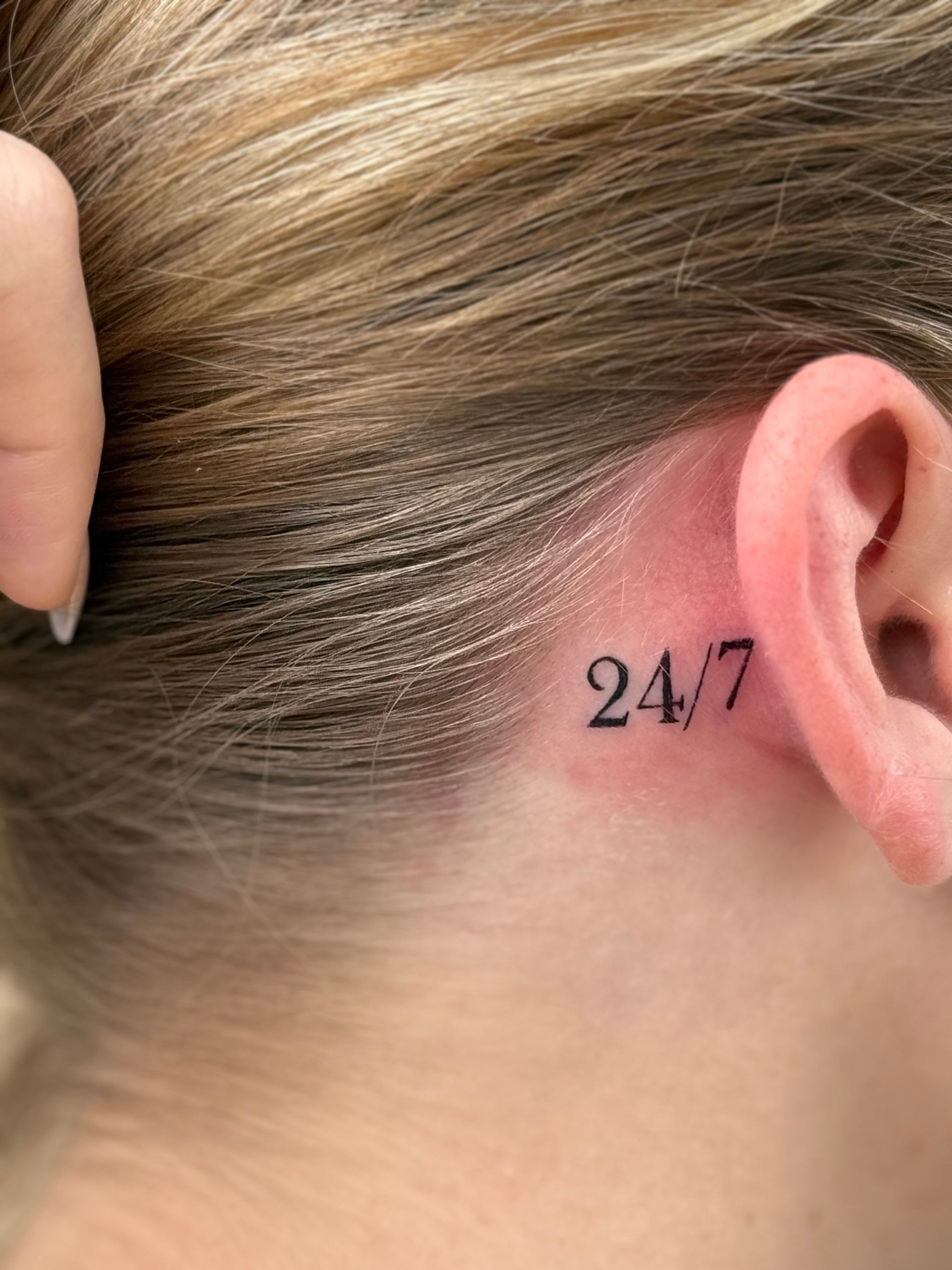Close-up of a person's ear and neck with a small 24/7 tattoo behind the ear