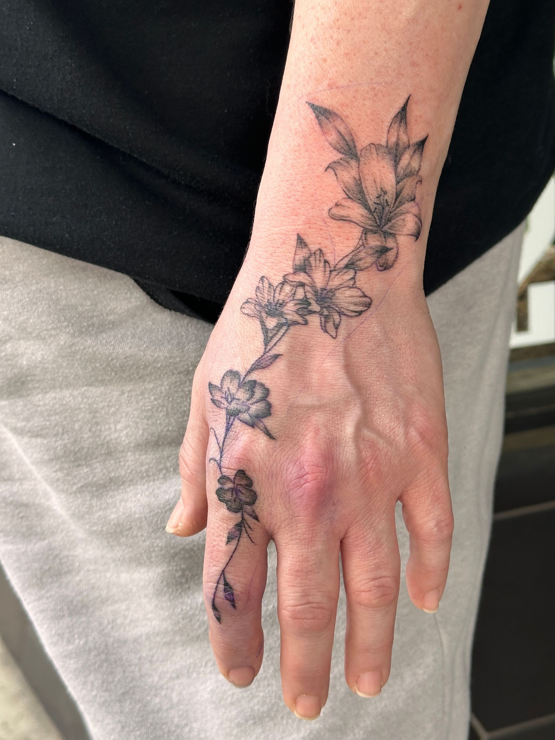 Hand with floral vine tattoo on wrist and fingers, resting against black and gray clothing.