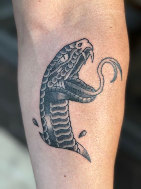 Black cobra tattoo on a forearm with its hood raised and tongue extended.