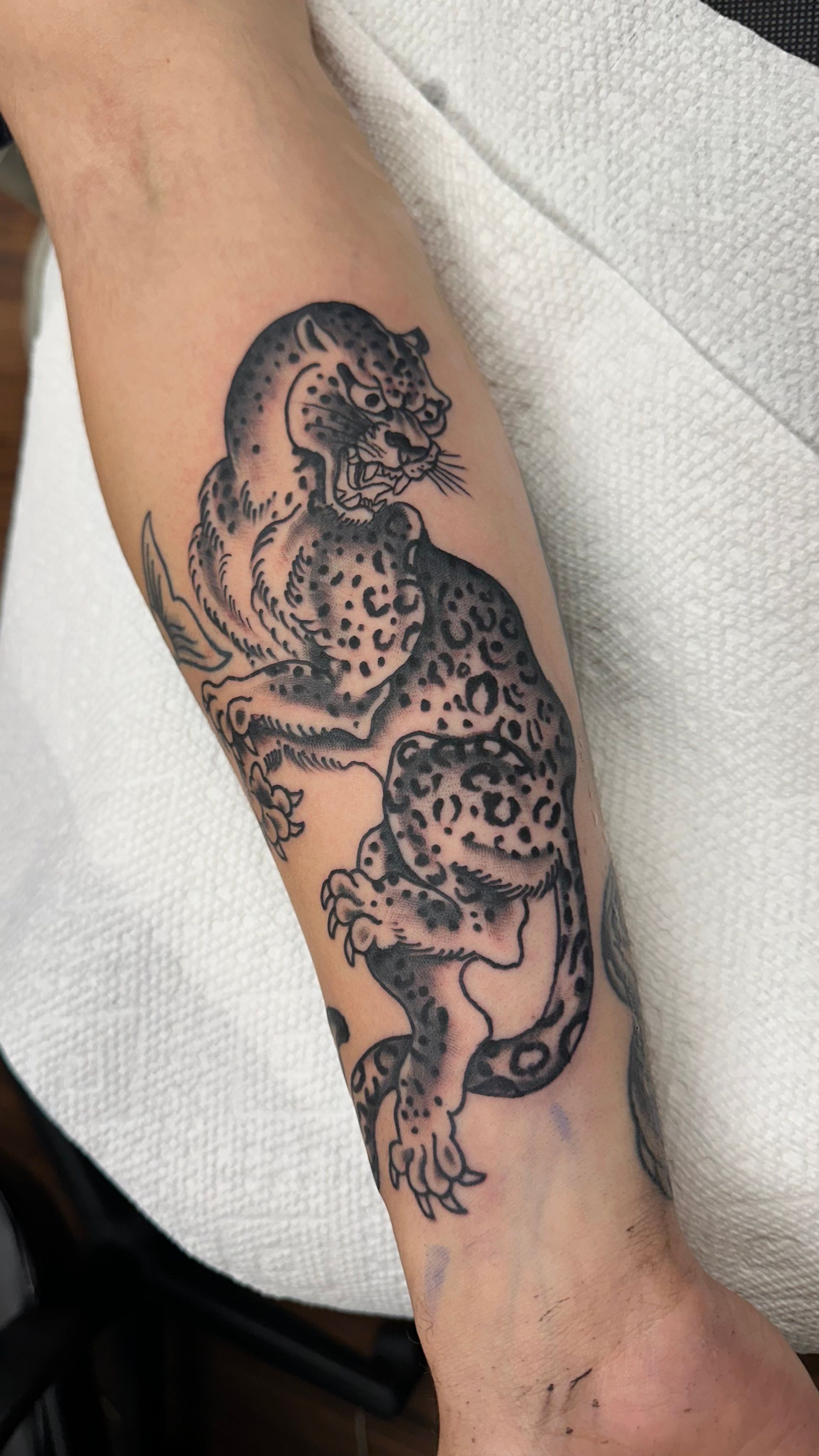 Black ink tattoo of a dragon-like creature on a person’s forearm, shown against a light fabric background.