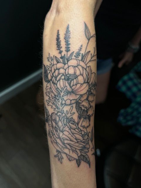 Black linework floral tattoo on a forearm with leaves and a large flower, shown close up.