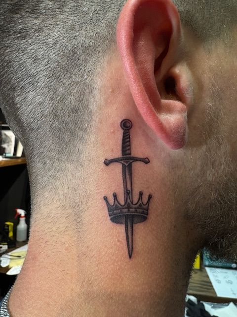 Sword and crown tattoo behind a shaved ear on the side of a neck