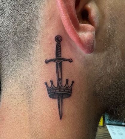 Sword and crown tattoo behind an ear on the side of a shaved head